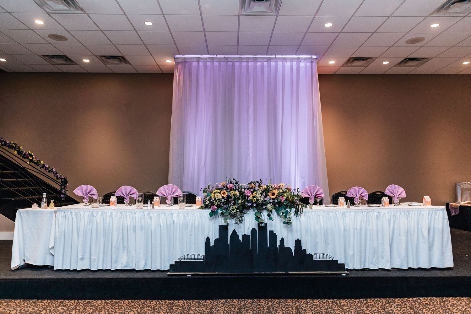 A wedding reception head table decorated with purple uplighting, white linens, and a city skyline silhouette backdrop.