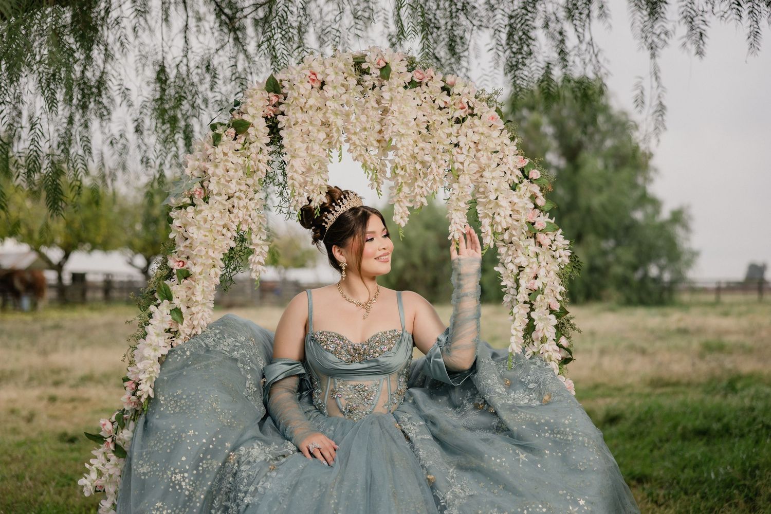 A quinceanera in a blue-gray tulle gown poses in front of an elegant floral arch in an outdoor garden setting during her photoshoot.