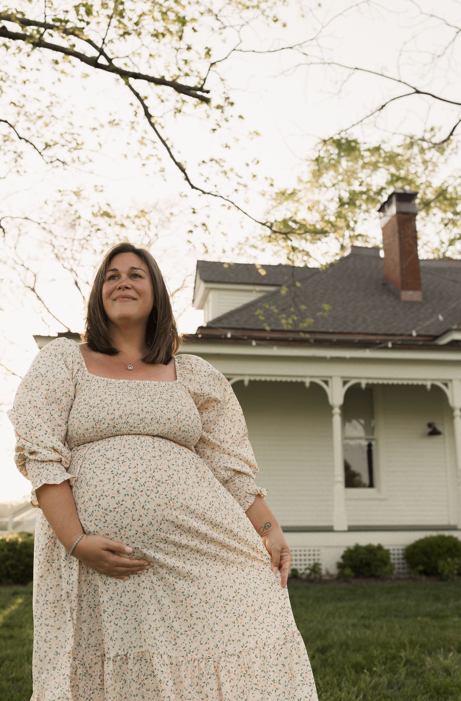 A springtime maternity portrait shows someone in a cream floral dress standing in front of a historic farmhouse.