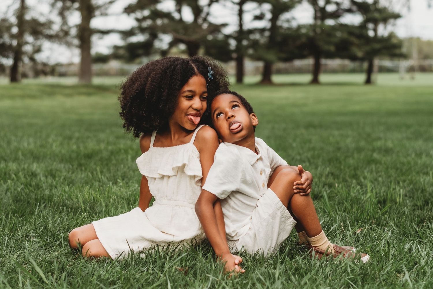 Two people in white outfits sit together laughing joyfully on green grass in a park setting.
