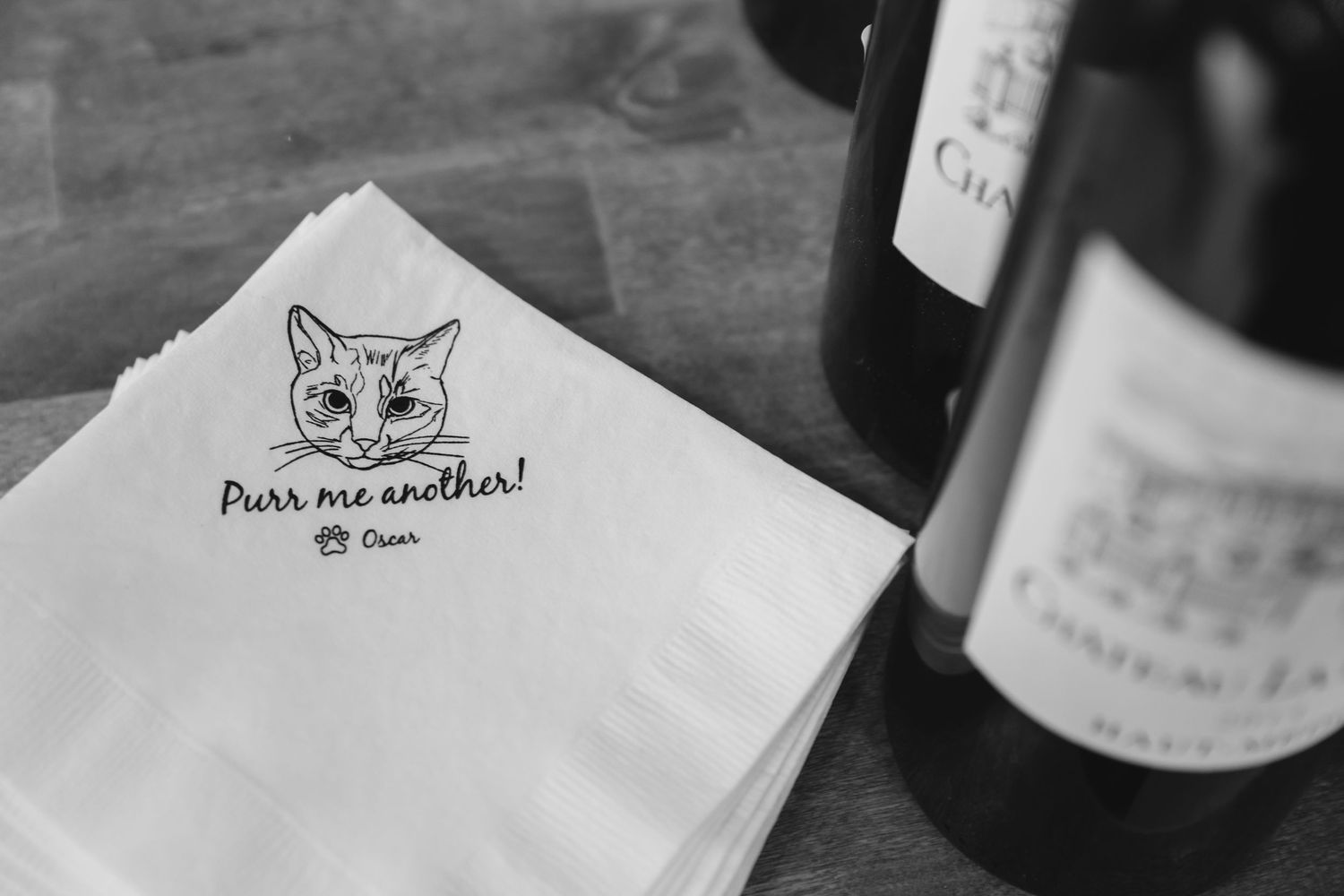 A white napkin with a cat drawing logo sits next to a wine bottle on a wooden table in black and white.
