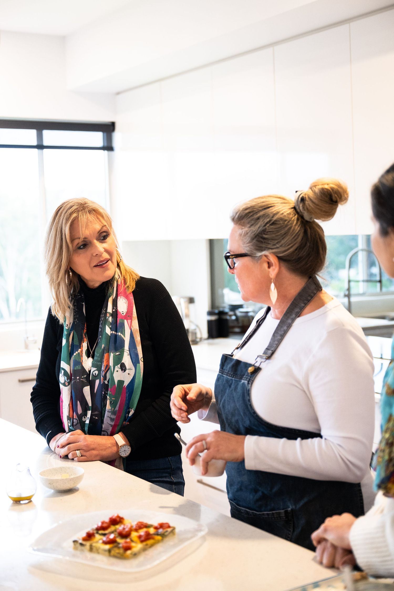 Two people having an animated conversation in a bright modern kitchen while preparing food.
