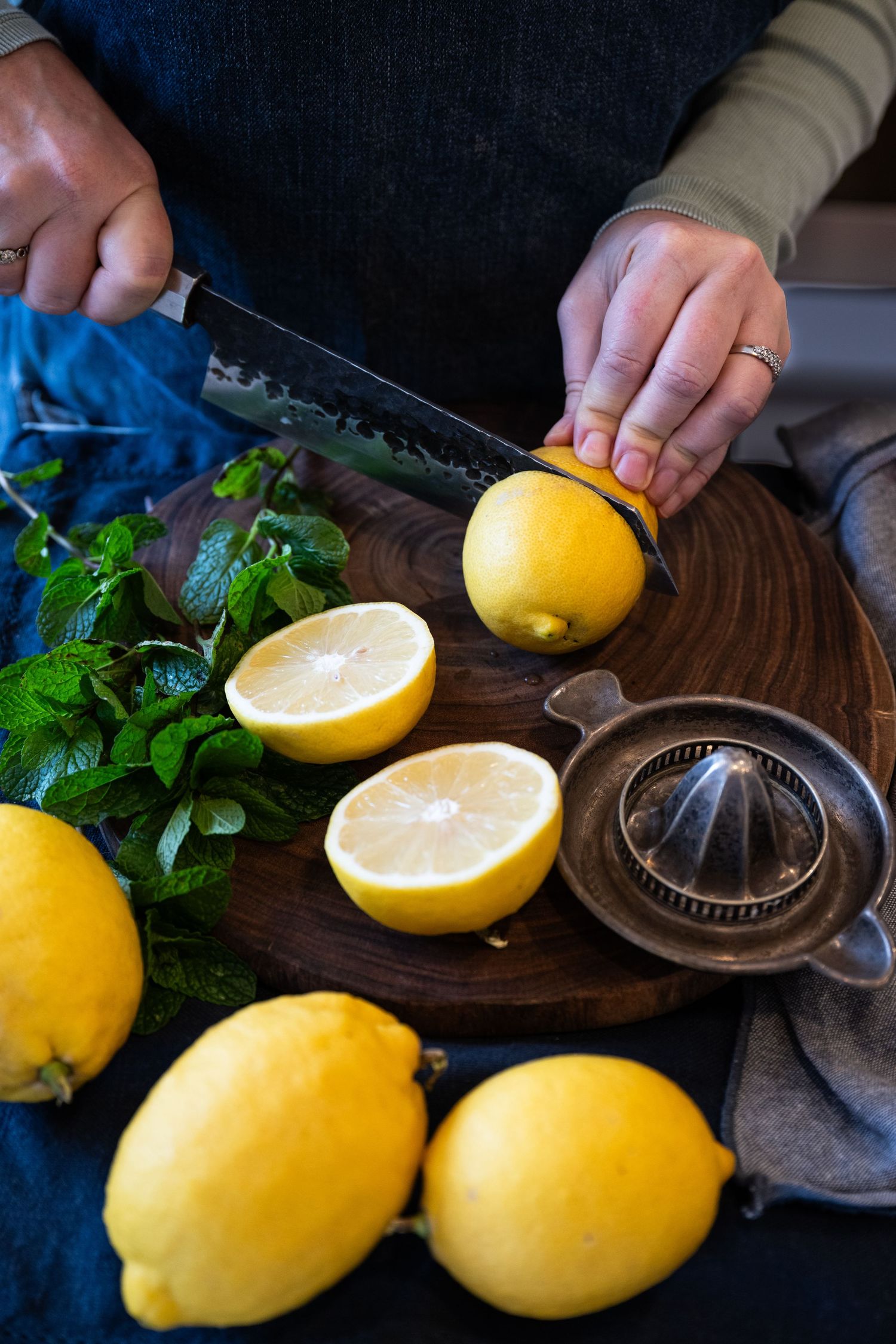 Fresh lemons and mint leaves on a wooden cutting board with a citrus juicer and hands slicing fruit.