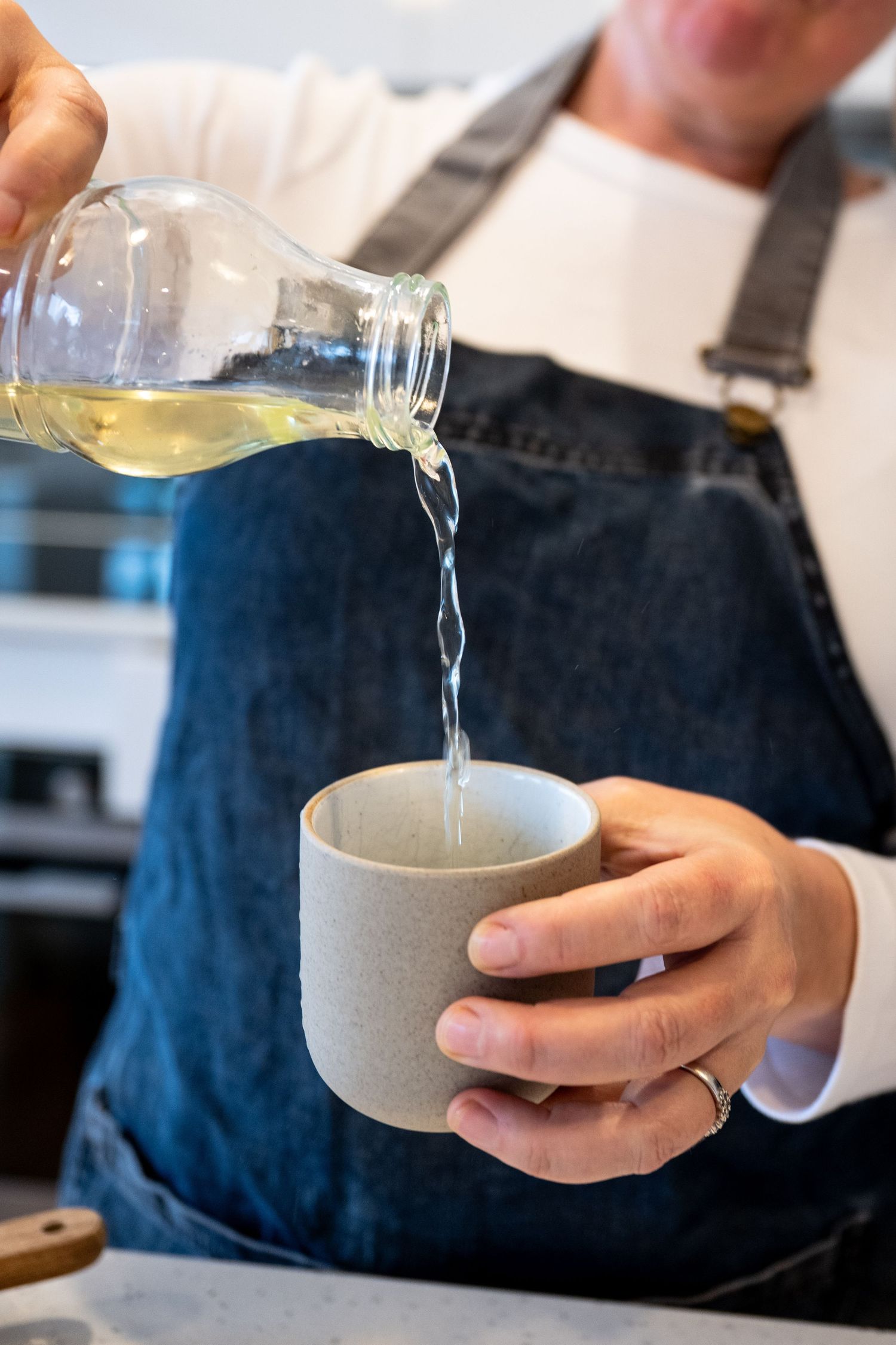 A person wearing a denim apron pours clear liquid from a glass bottle into a gray ceramic cup.