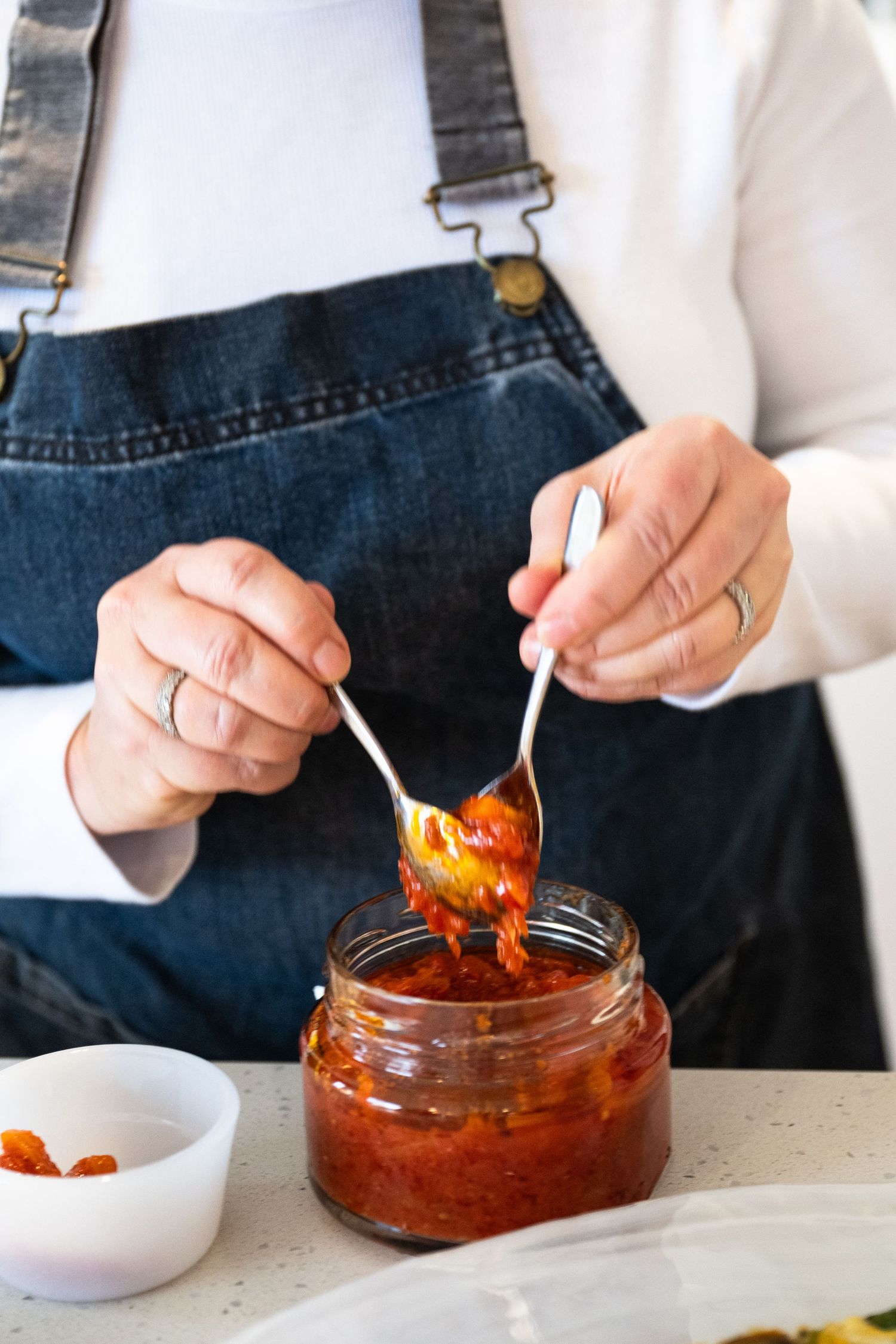 A hand with a spoon scooping out vibrant red chili oil or hot sauce from a glass jar on a white countertop.