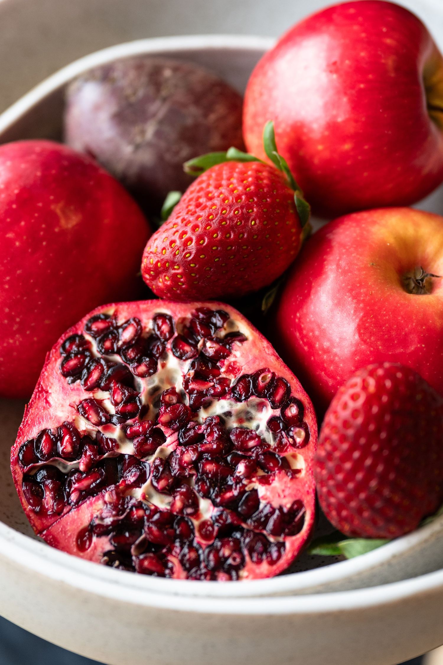 Fresh red fruits including pomegranate, apples and strawberries arranged in a white ceramic bowl.
