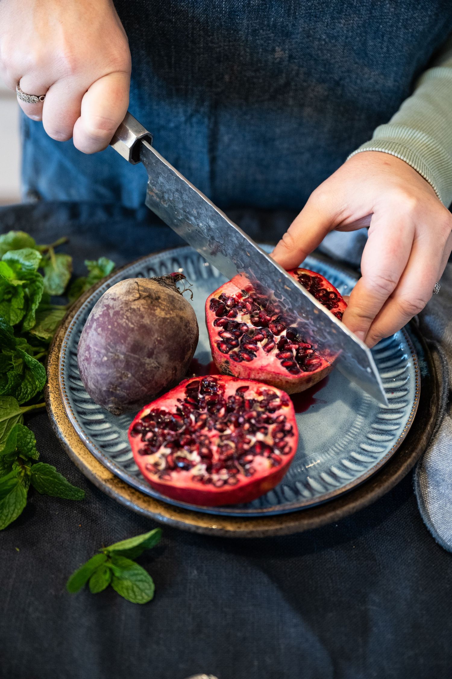 Someone cutting fresh pomegranates on a dark ceramic plate with mint leaves scattered around.