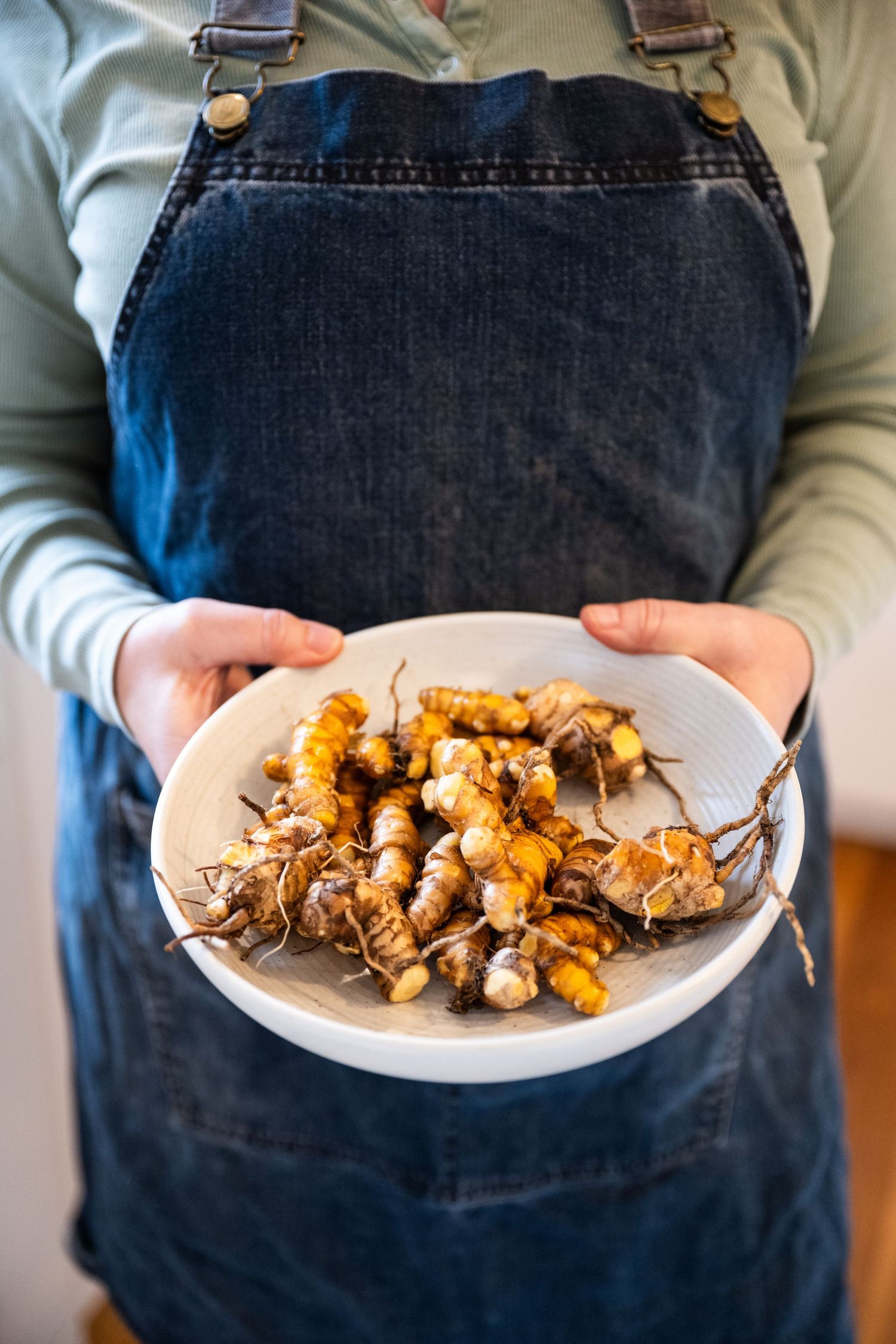 Someone wearing denim overalls holds a plate of freshly foraged wild mushrooms and turmeric root.