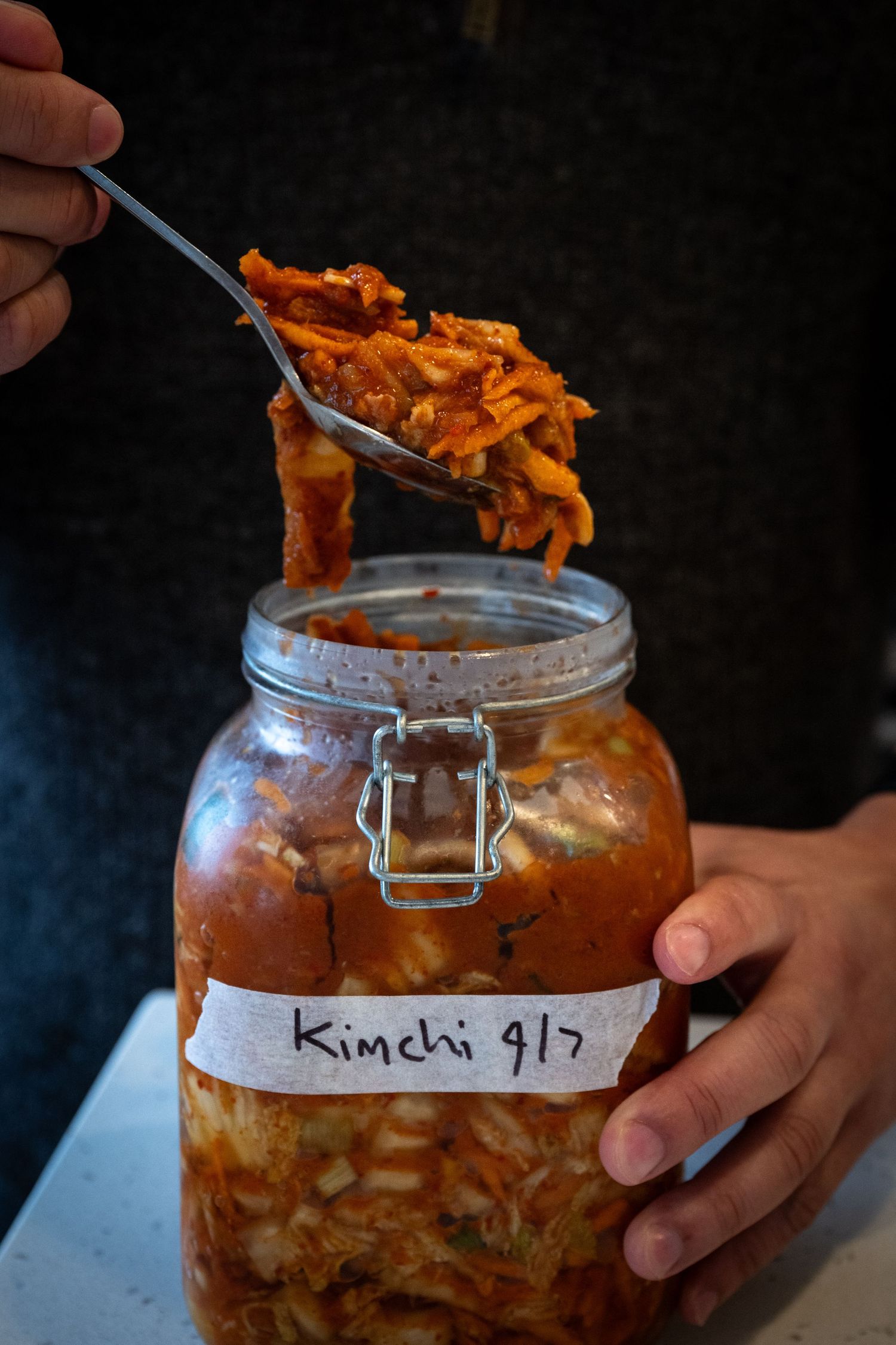 A jar of homemade kimchi with a spoon lifting out spicy fermented cabbage against a dark background.