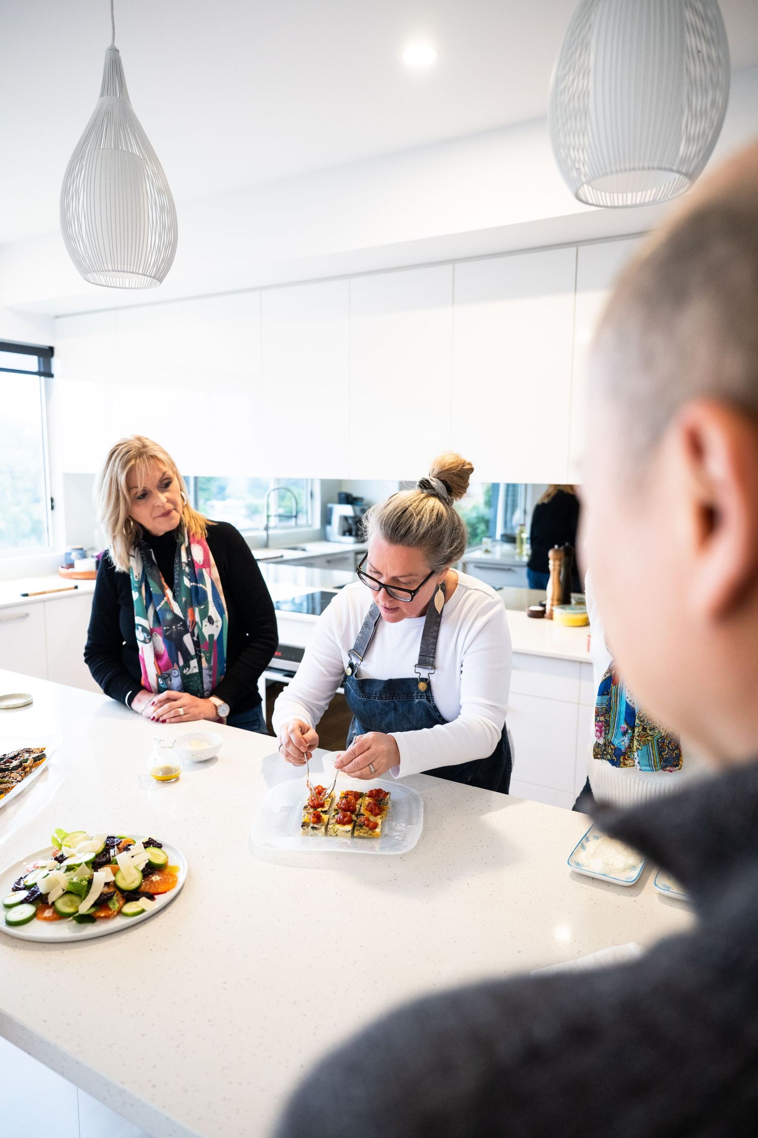 People gather at a sleek modern kitchen counter with white pendant lights while preparing and plating food together.
