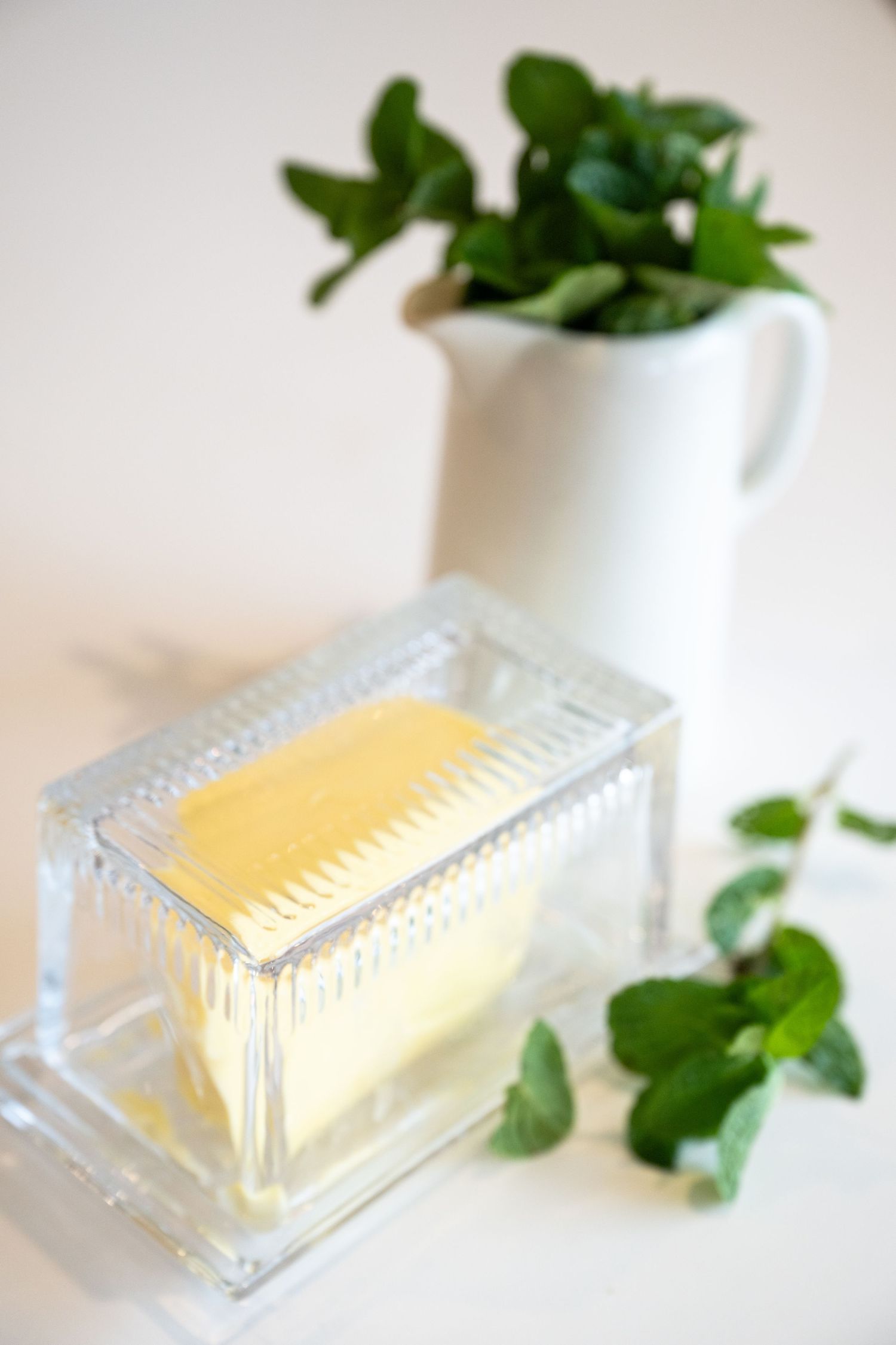 A stick of butter in a glass dish with fresh green herbs in a white ceramic pitcher against a light background.