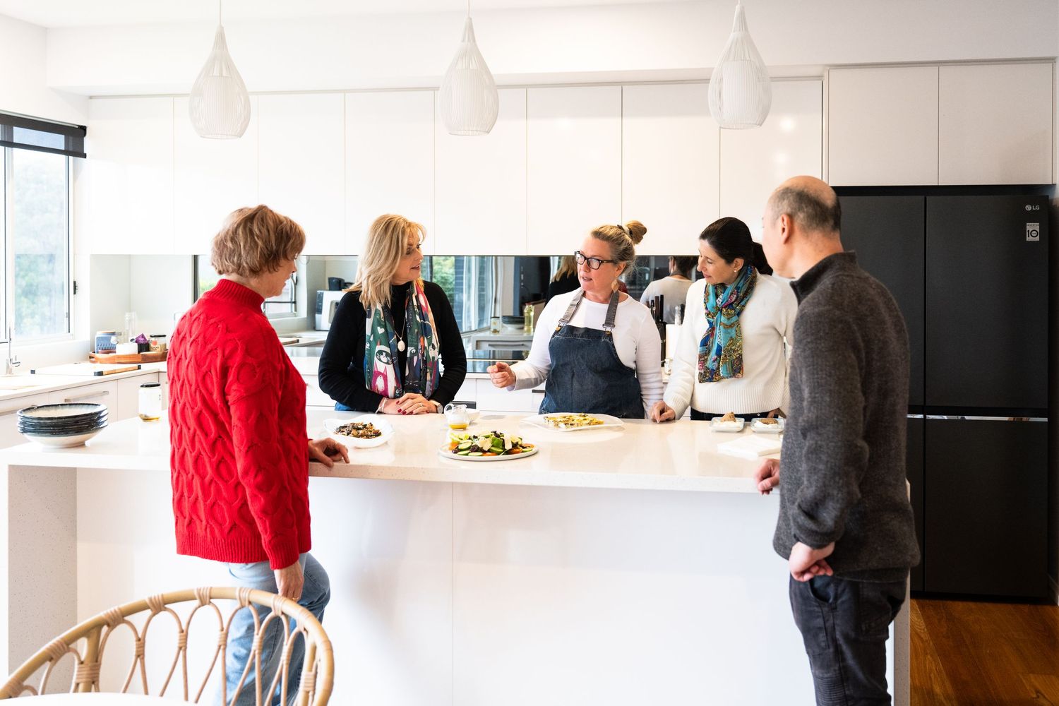 Group of people gathered in a modern white kitchen space around counter for food preparation or cooking class.