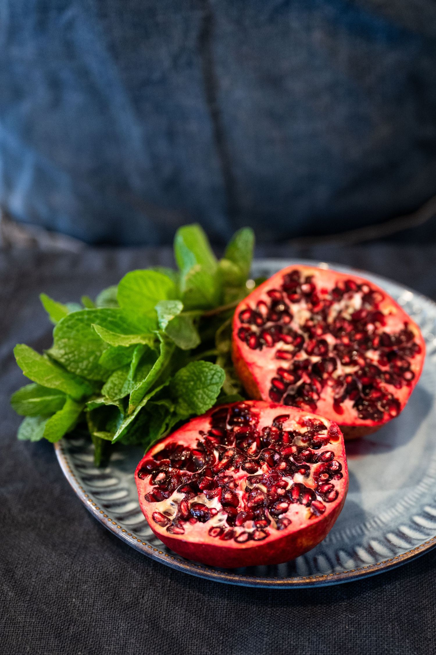 Fresh halved pomegranate with bright red seeds and mint leaves on a blue plate against dark background.
