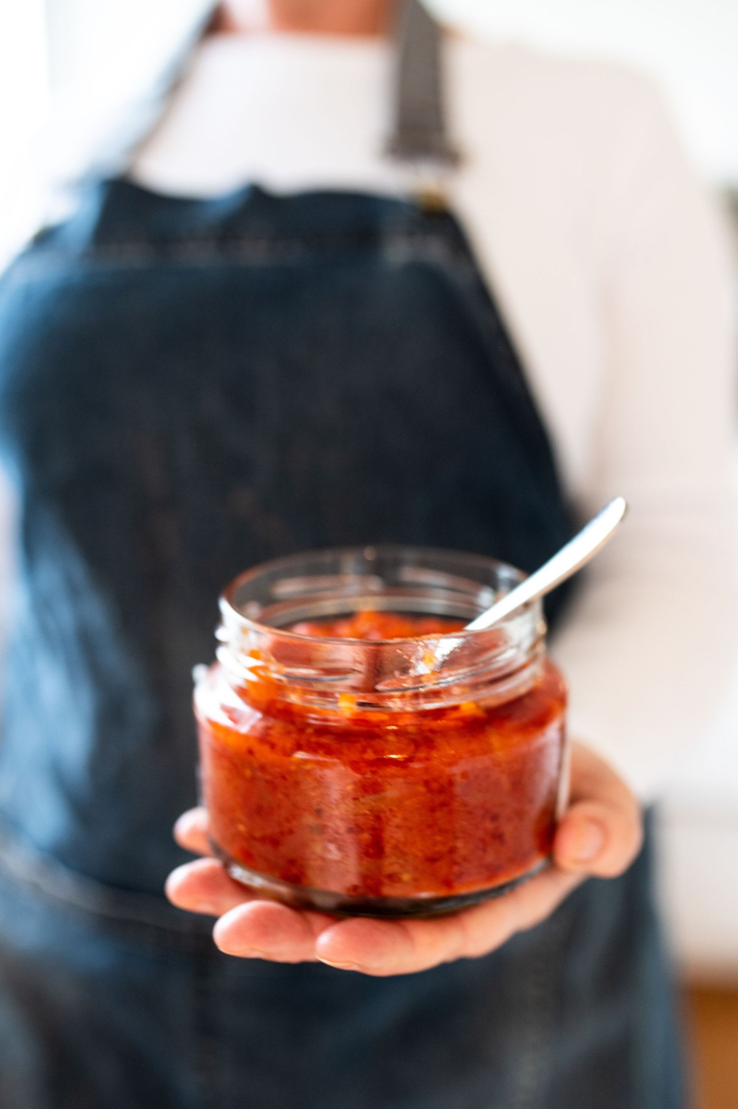 Hands hold a glass jar containing vibrant red harissa sauce or spicy chili paste against a blurred background.