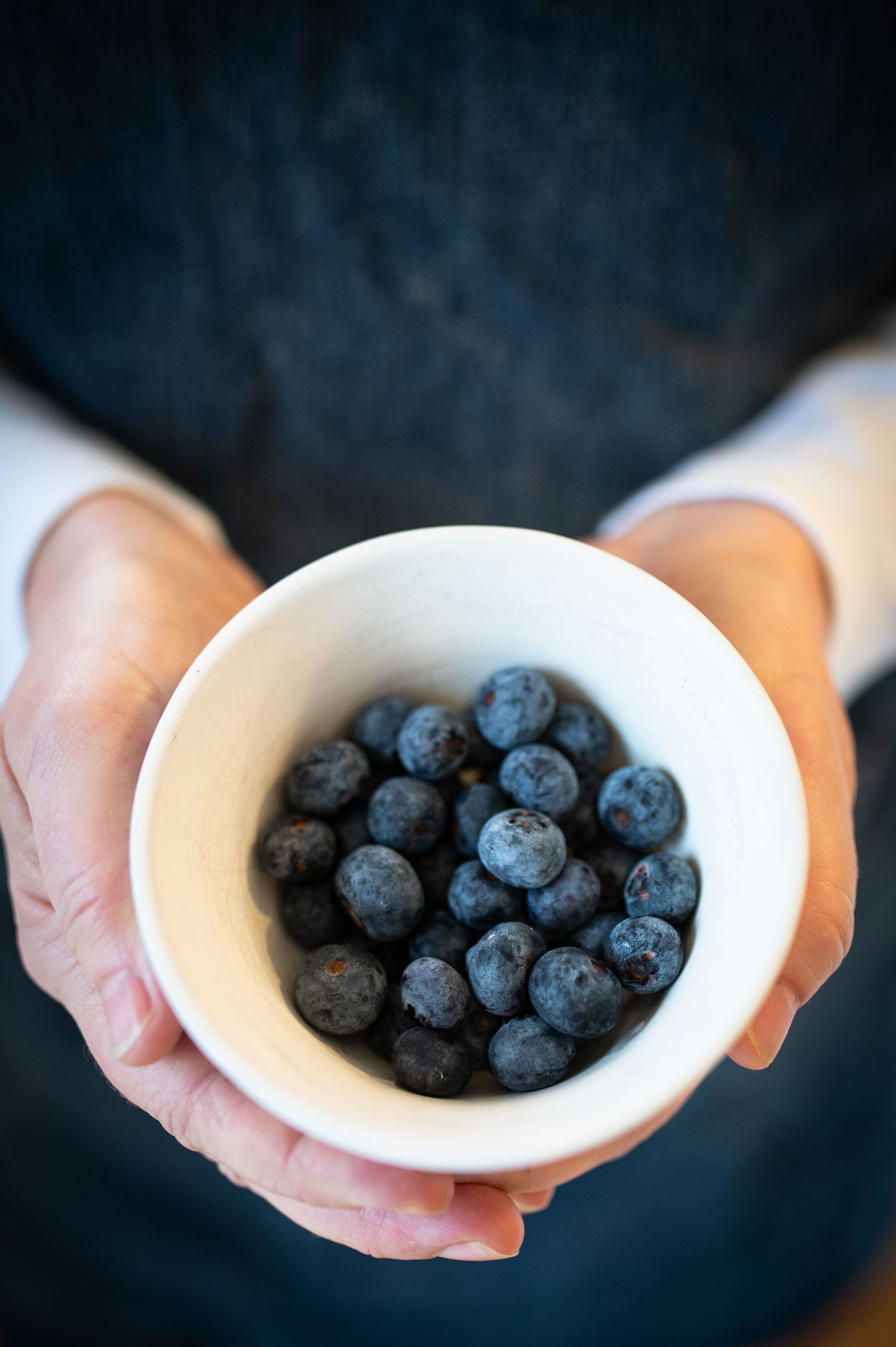 Hands holding a small white ceramic bowl filled with fresh ripe blueberries against a dark background.
