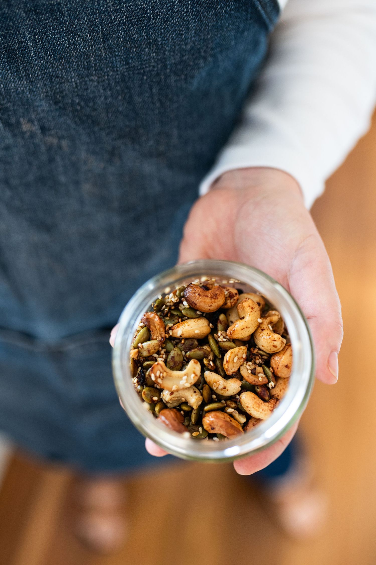 Hand holding glass jar filled with seasoned mixed nuts viewed from above on wooden floor.