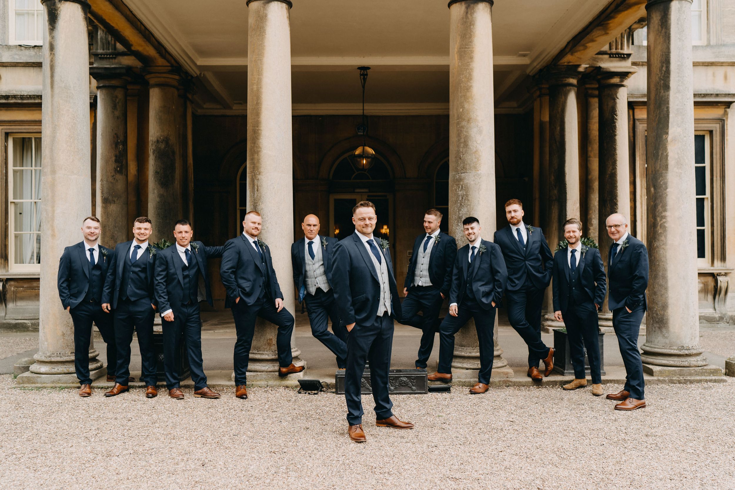 Groomsman in matching navy suits poses together on grand columned porch of historic building of Prestwold Hall.