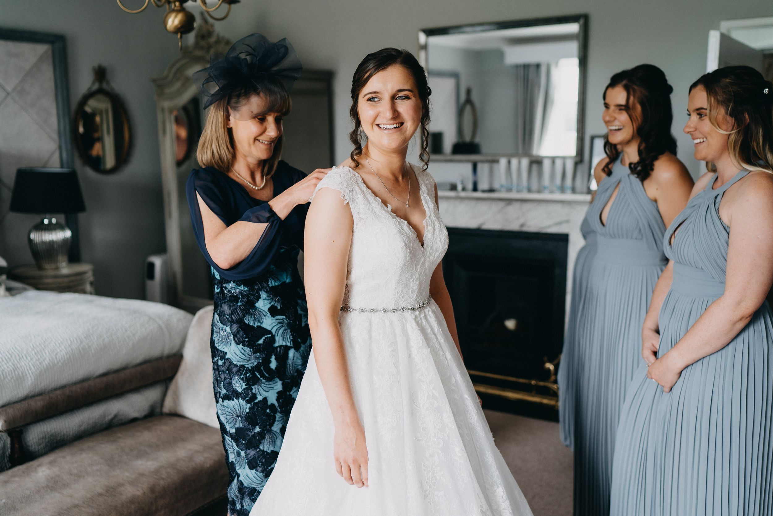 A glowing bride in a white lace gown shares a joyful moment with bridesmaids in blue dresses during wedding preparations.
