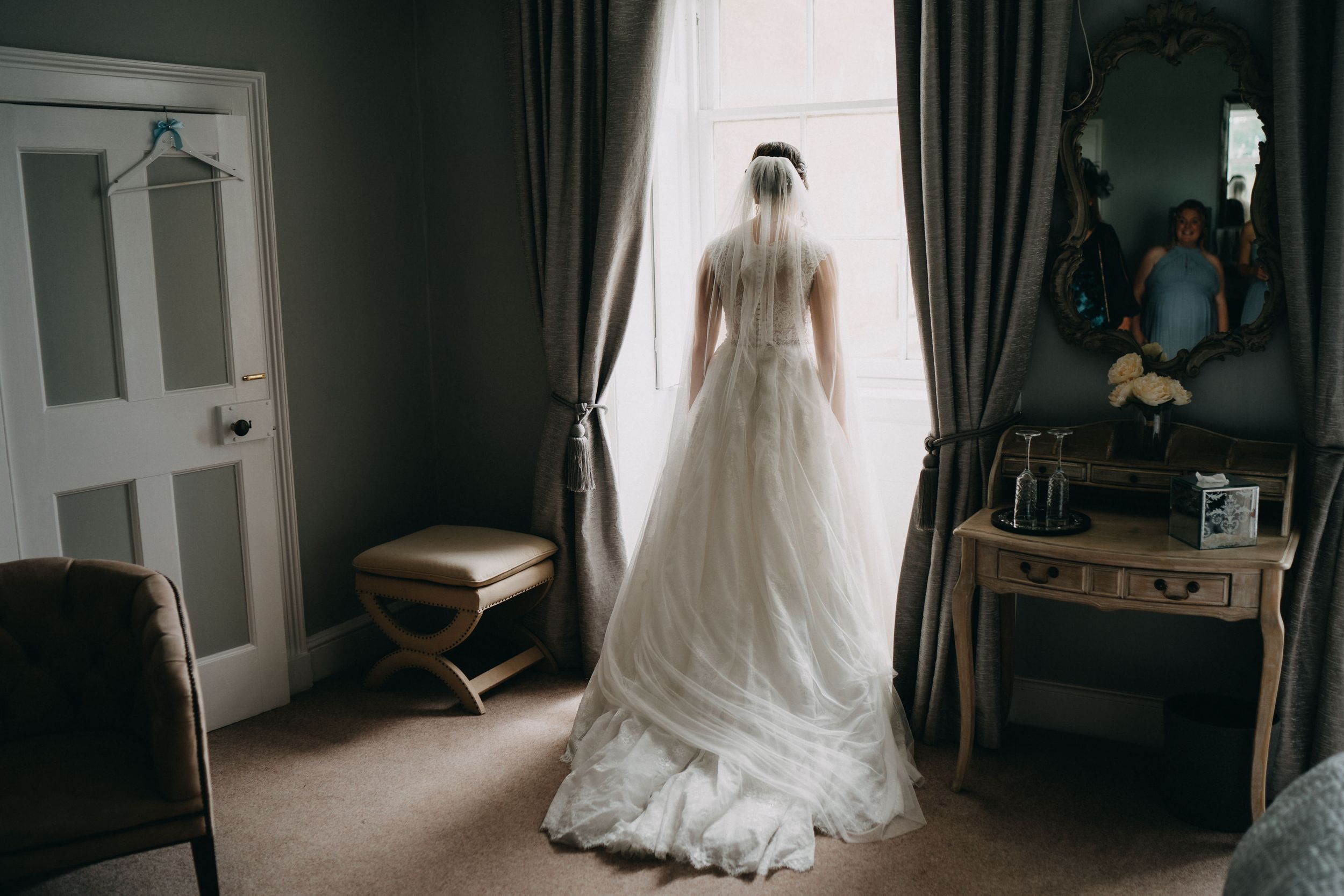 A bride in an elegant white wedding gown stands silhouetted against a window in a softly lit vintage bedroom.