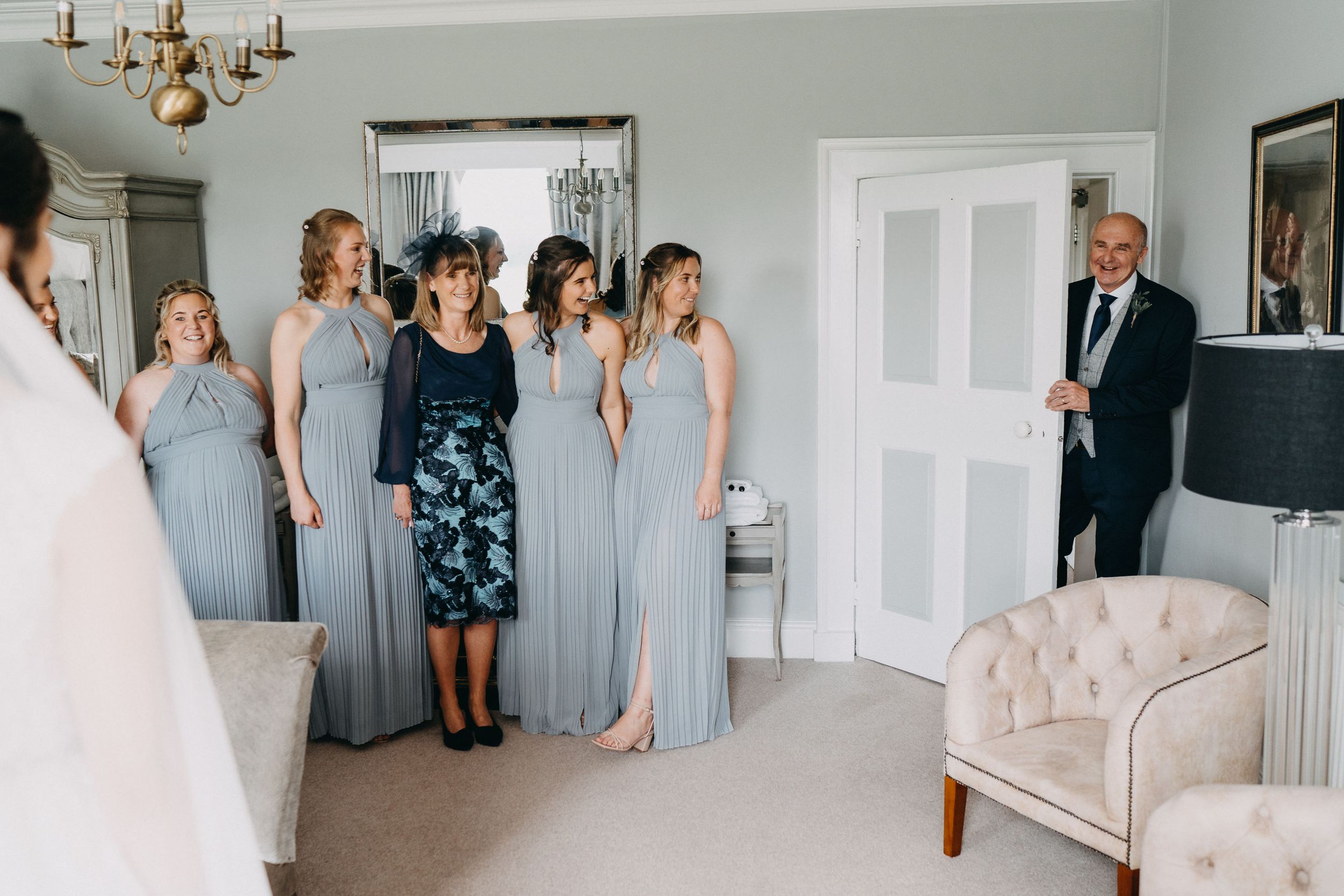People in pale blue bridesmaid dresses gather in an elegant room with white doors and a chandelier during wedding preparations.