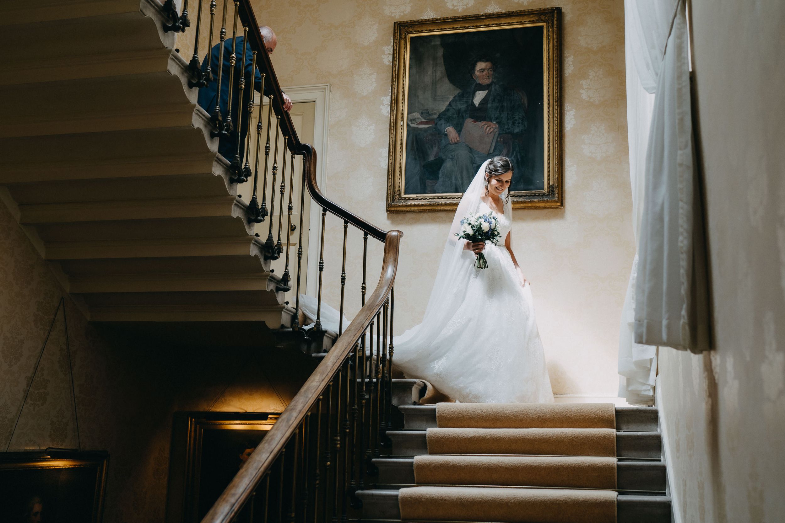 Elegant bride in white wedding dress poses on grand staircase at Prestwold Hall next to vintage portrait in historic mansion interior.