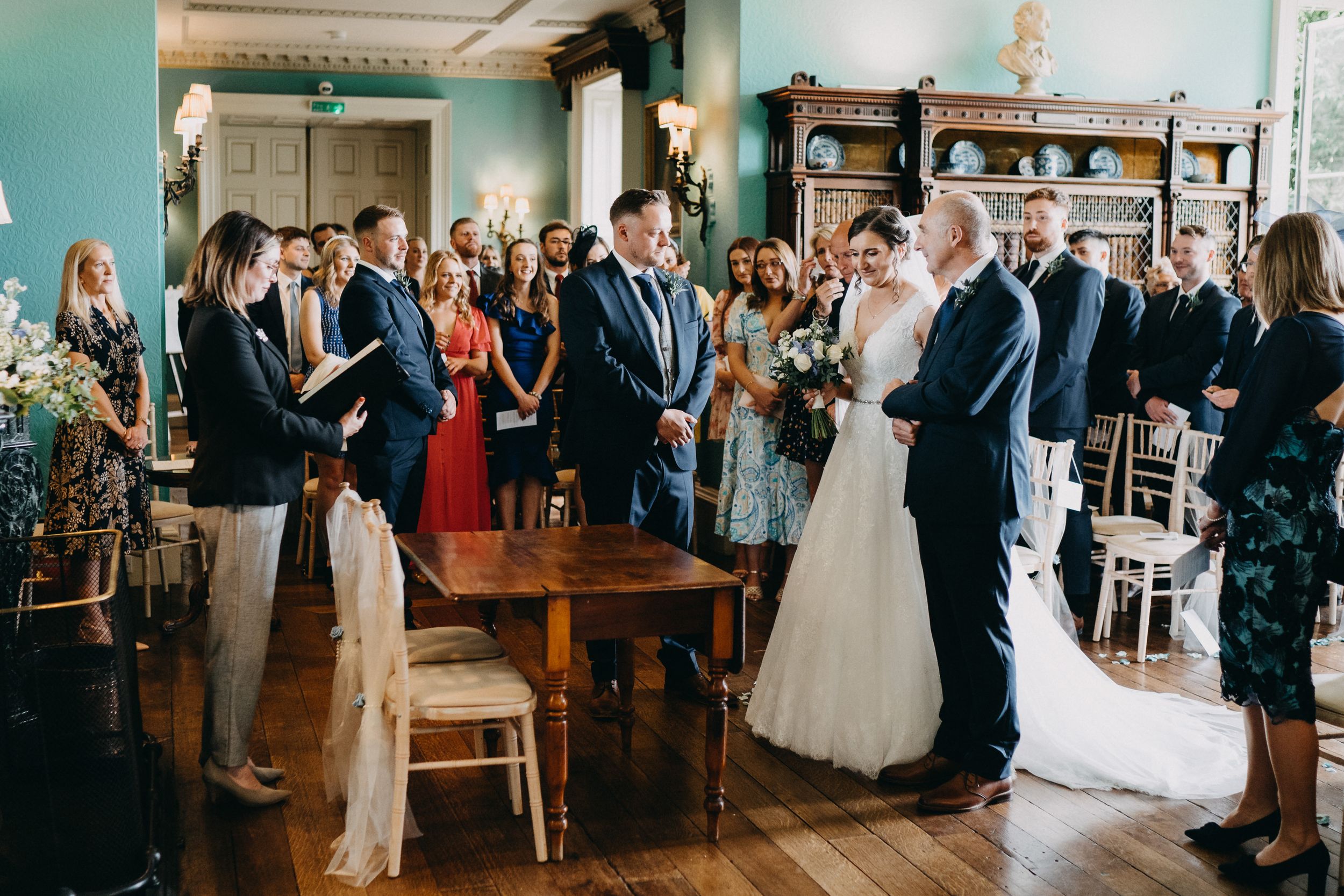 Wedding guests gather in a vintage-style room with turquoise walls as the ceremony unfolds at the front of the space.