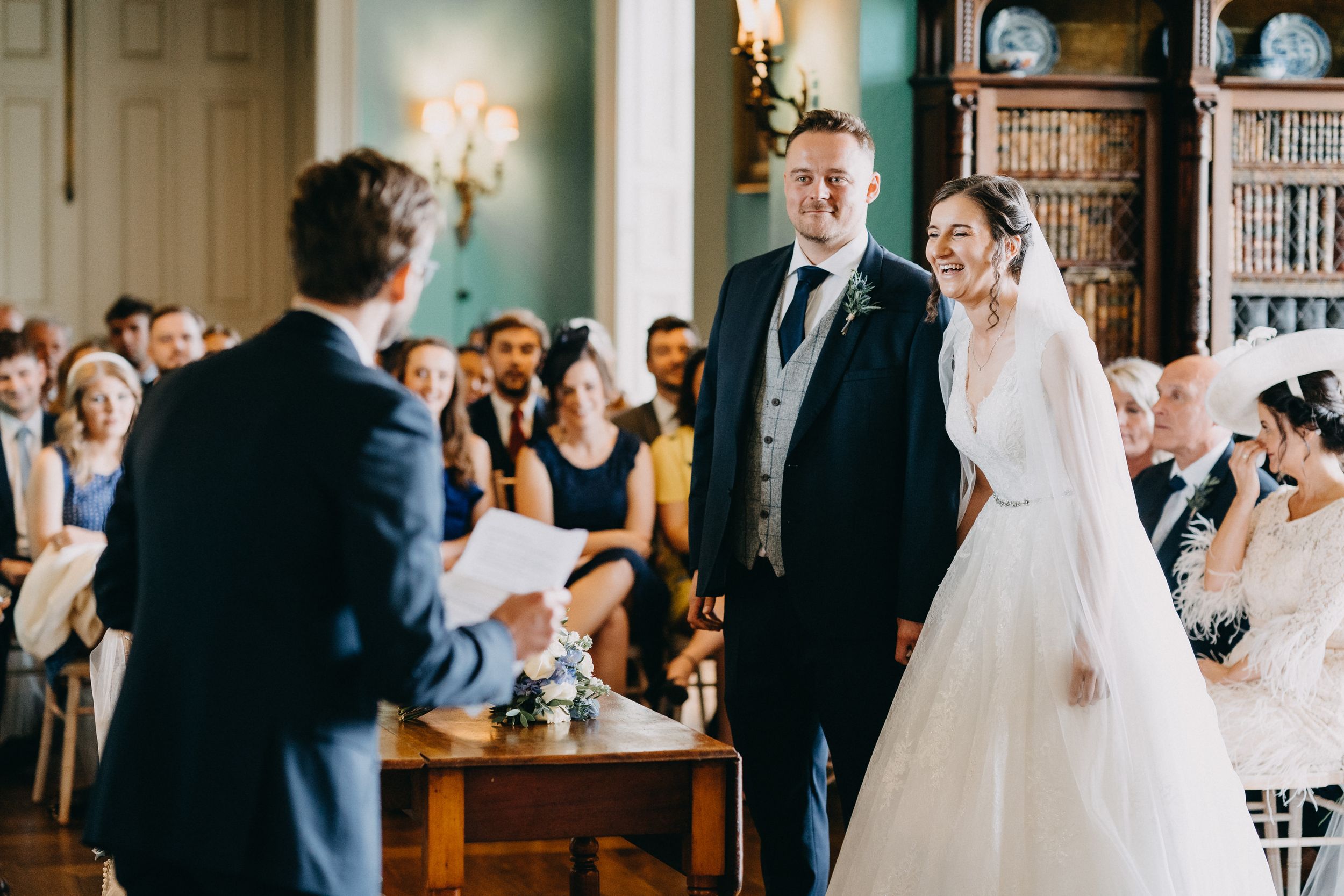 Traditional wedding ceremony at Prestwold Hall takes place in ornate room with bookshelf-lined walls and glowing wall sconces.