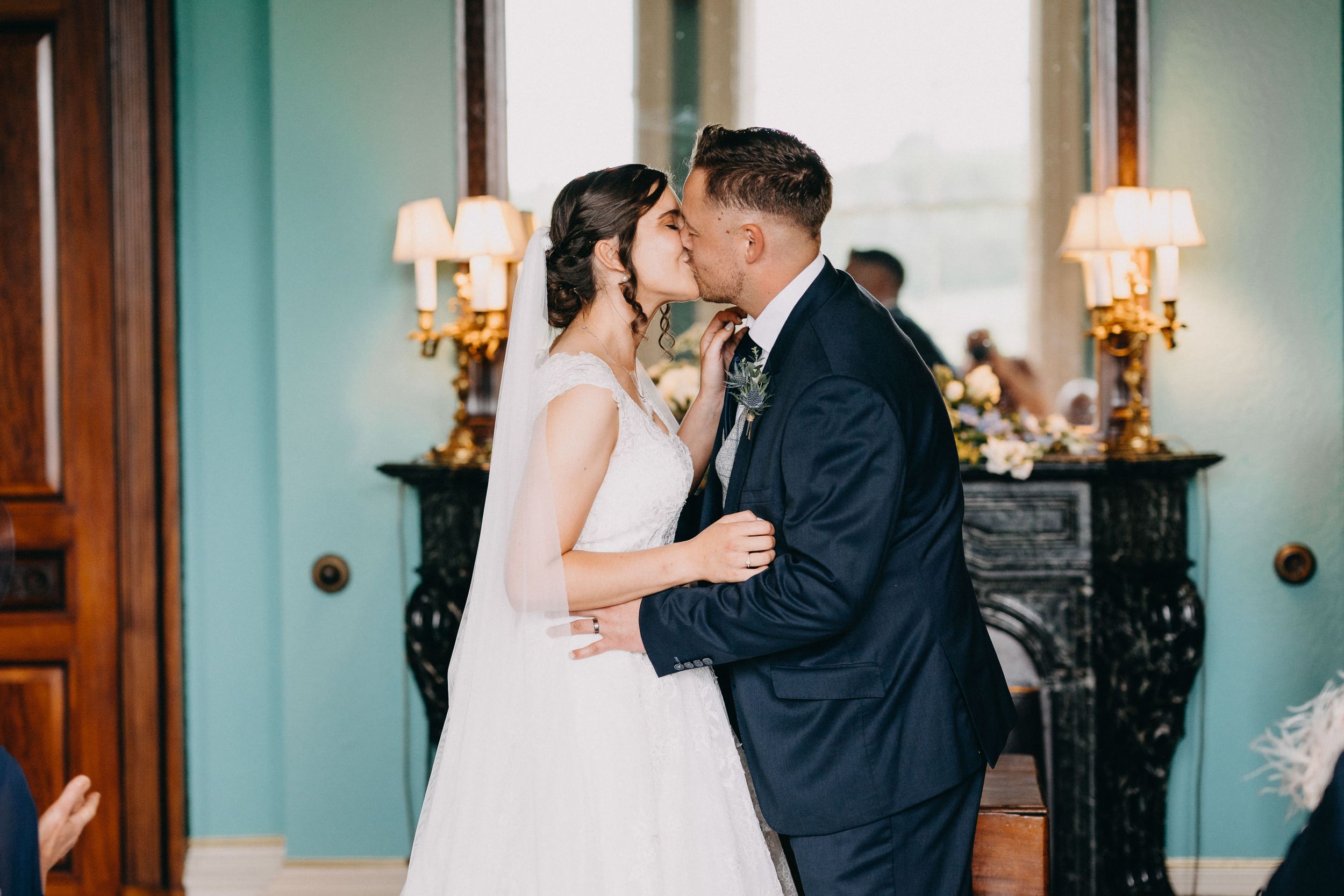 A romantic wedding moment with the couple sharing a tender kiss in front of ornate wall sconces and decor.