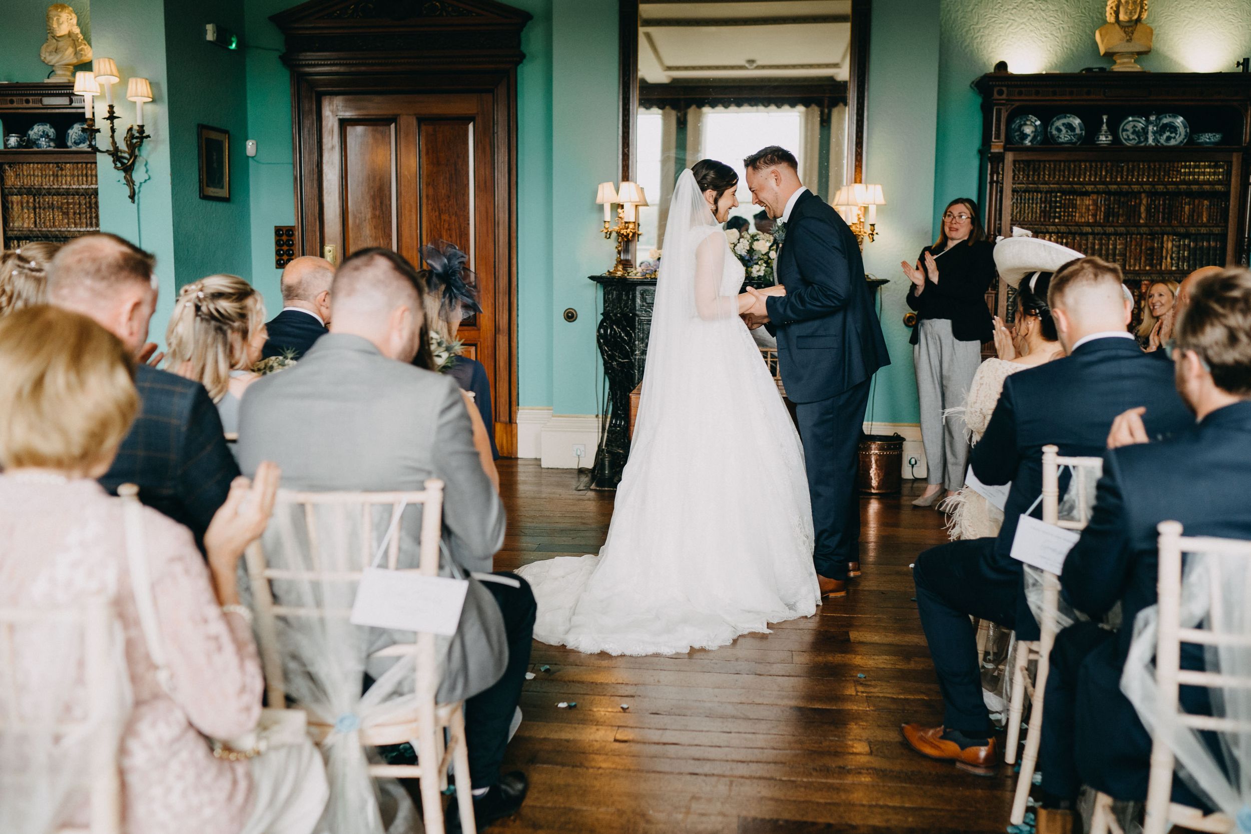 Indoor wedding ceremony with guests seated in white chairs inside an elegant green-walled room with vintage decor.