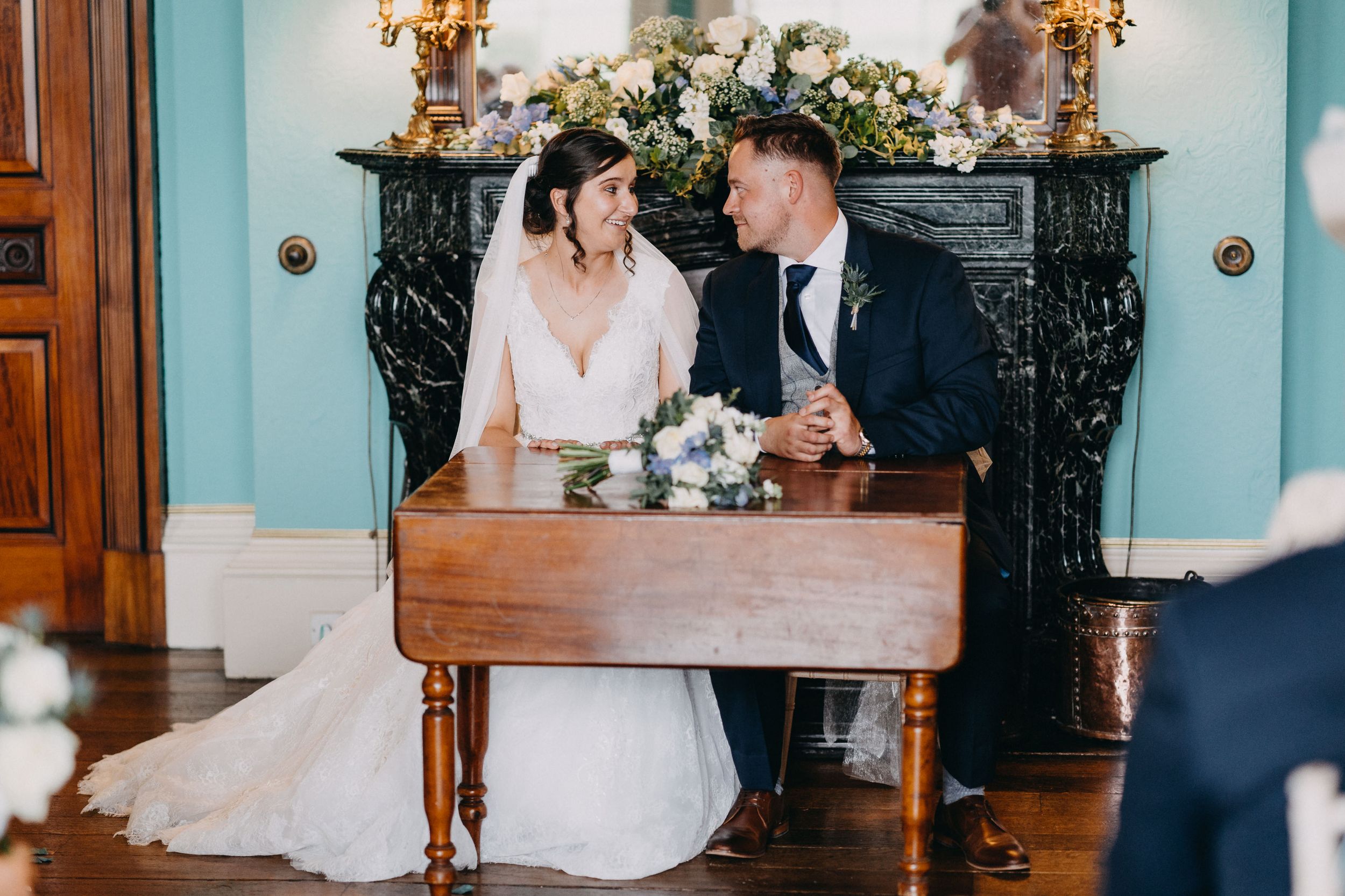 Wedding couple shares an intimate moment while sitting at an antique wooden table against a turquoise wall with marble fireplace.