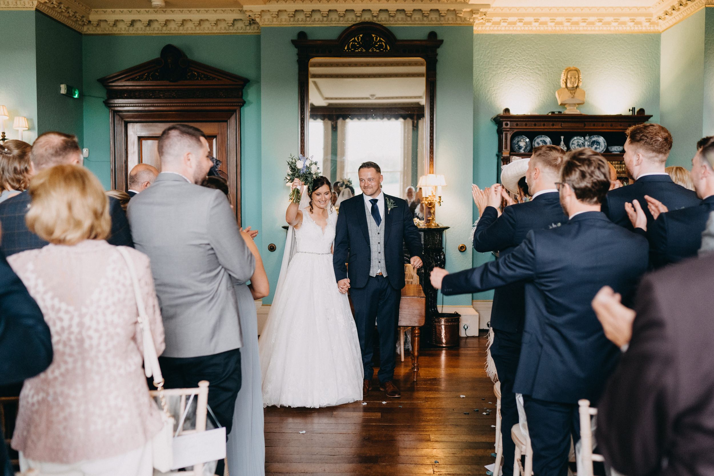 Wedding guests stand along the aisle in an elegant teal-walled room with ornate molding as a couple makes their entrance.