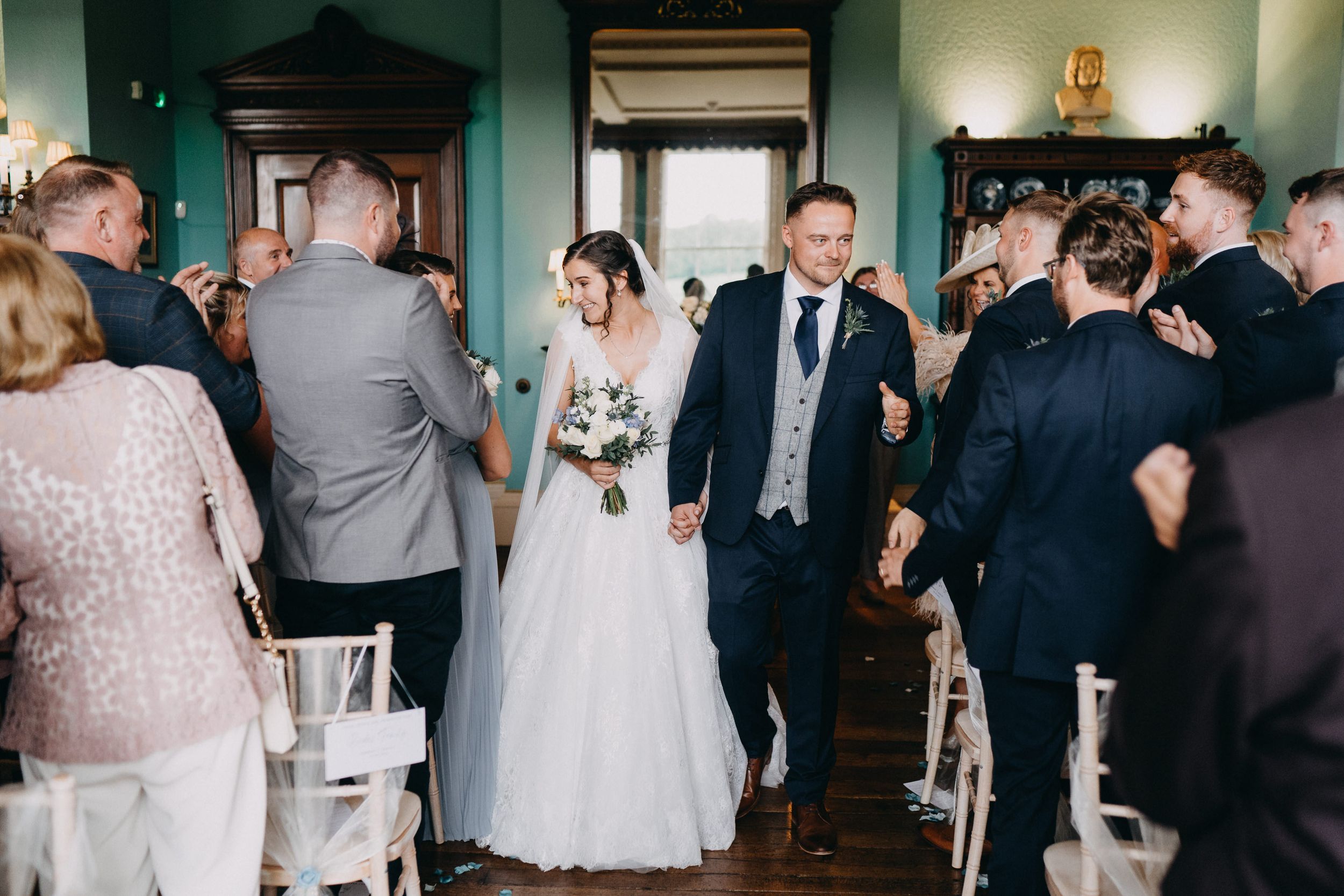 A wedding ceremony celebration at an indoor venue with guests lining the aisle as the bride walks through in a white gown.