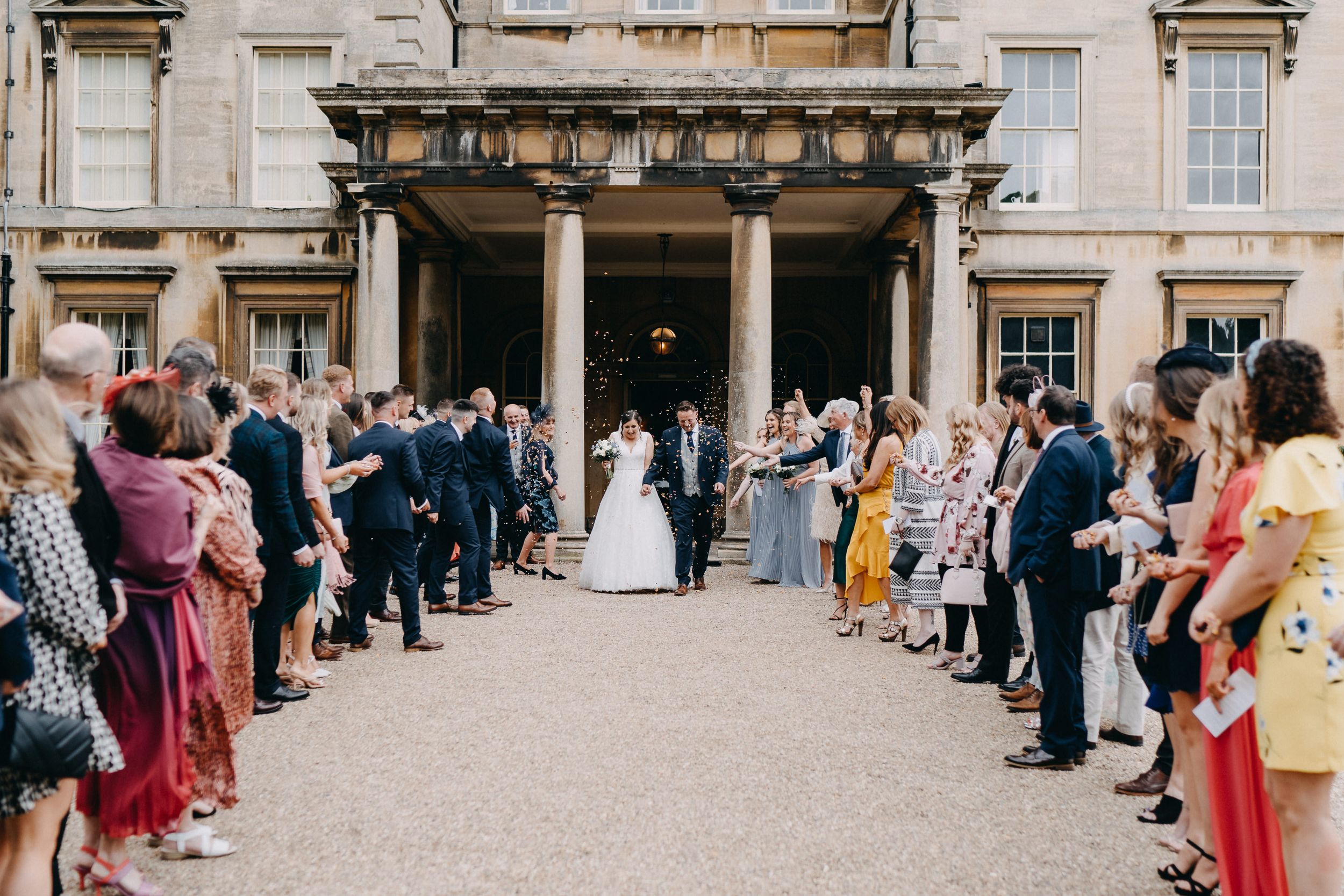 Wedding guests line up outside a grand historic building of Prestwold Hall as a couple exits through ornate entrance doors.