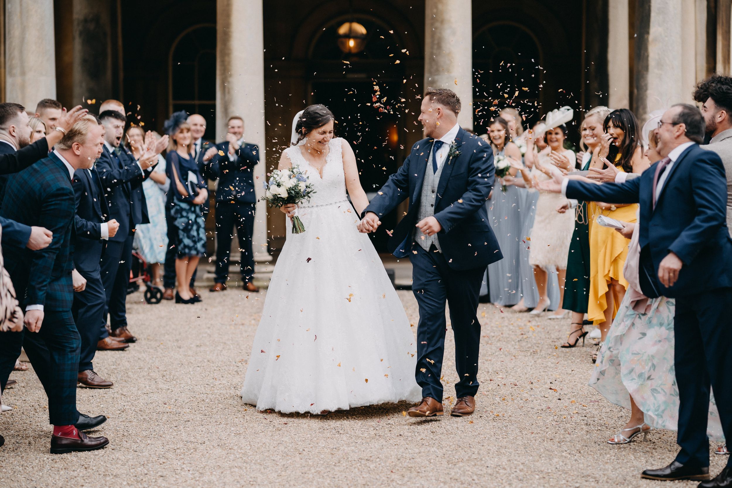 Wedding guests throw confetti on newlyweds during outdoor celebration while crowd watches along pathway.