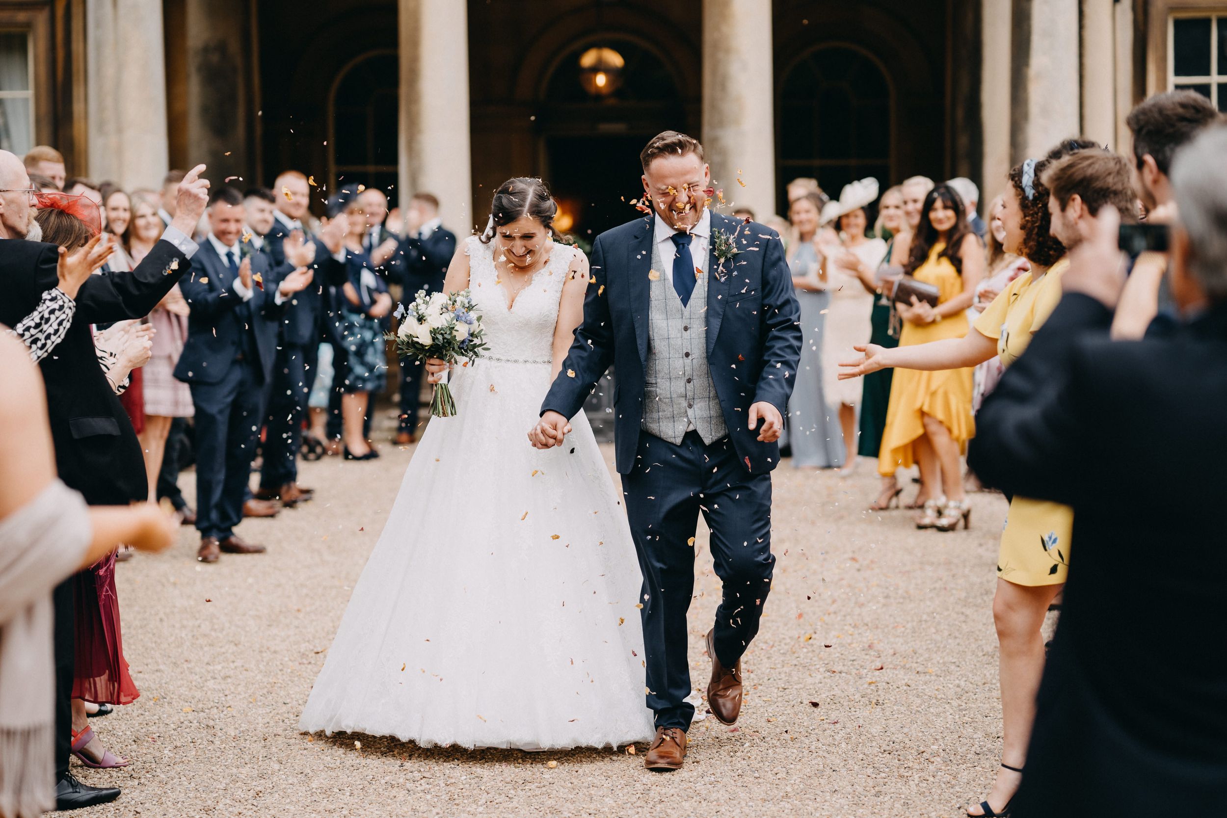 A newlywed couple runs joyfully through a crowd of guests throwing confetti at a grand venue entrance.