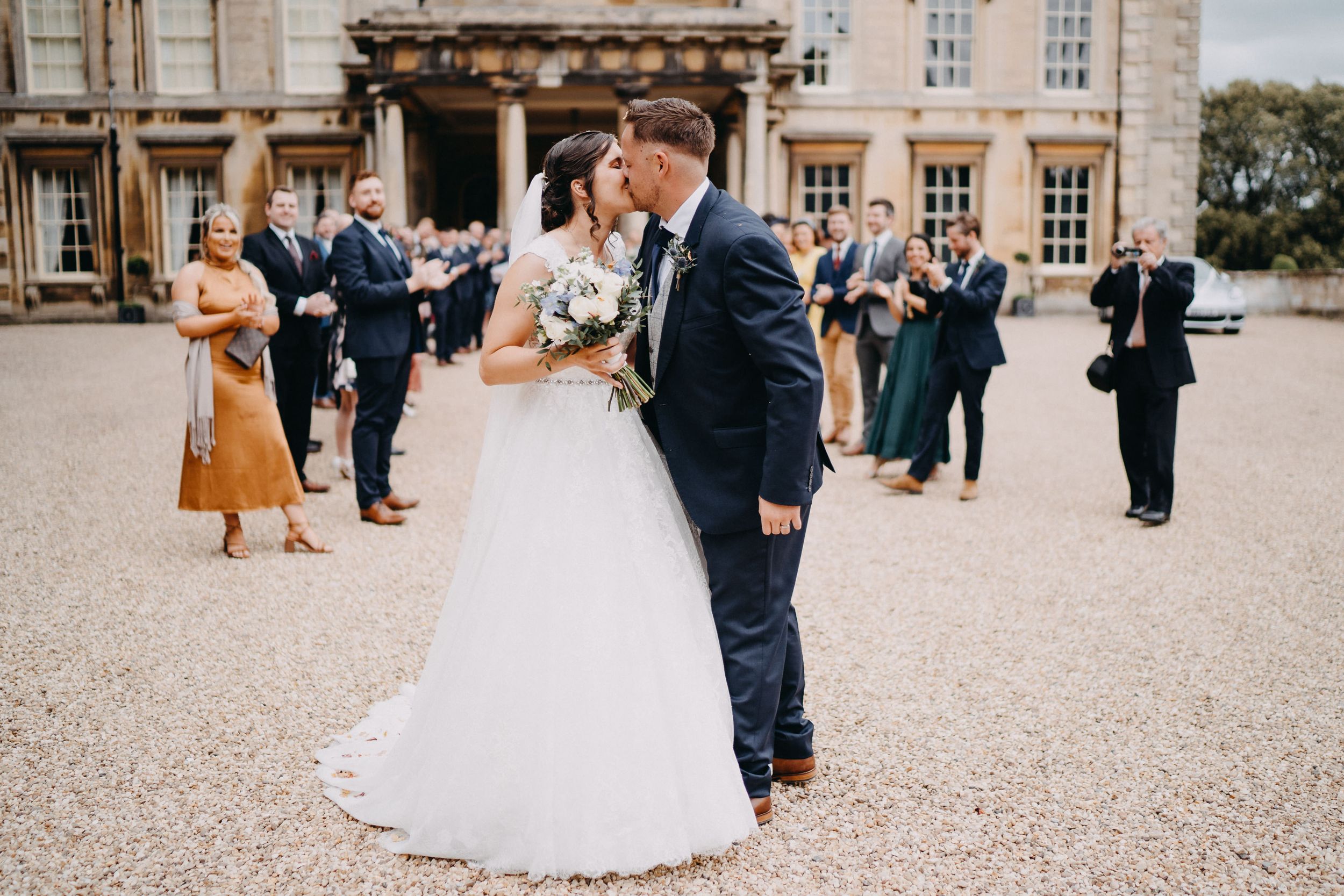 A couple sharing a tender moment in wedding attire outside a grand historic building with guests in the background.