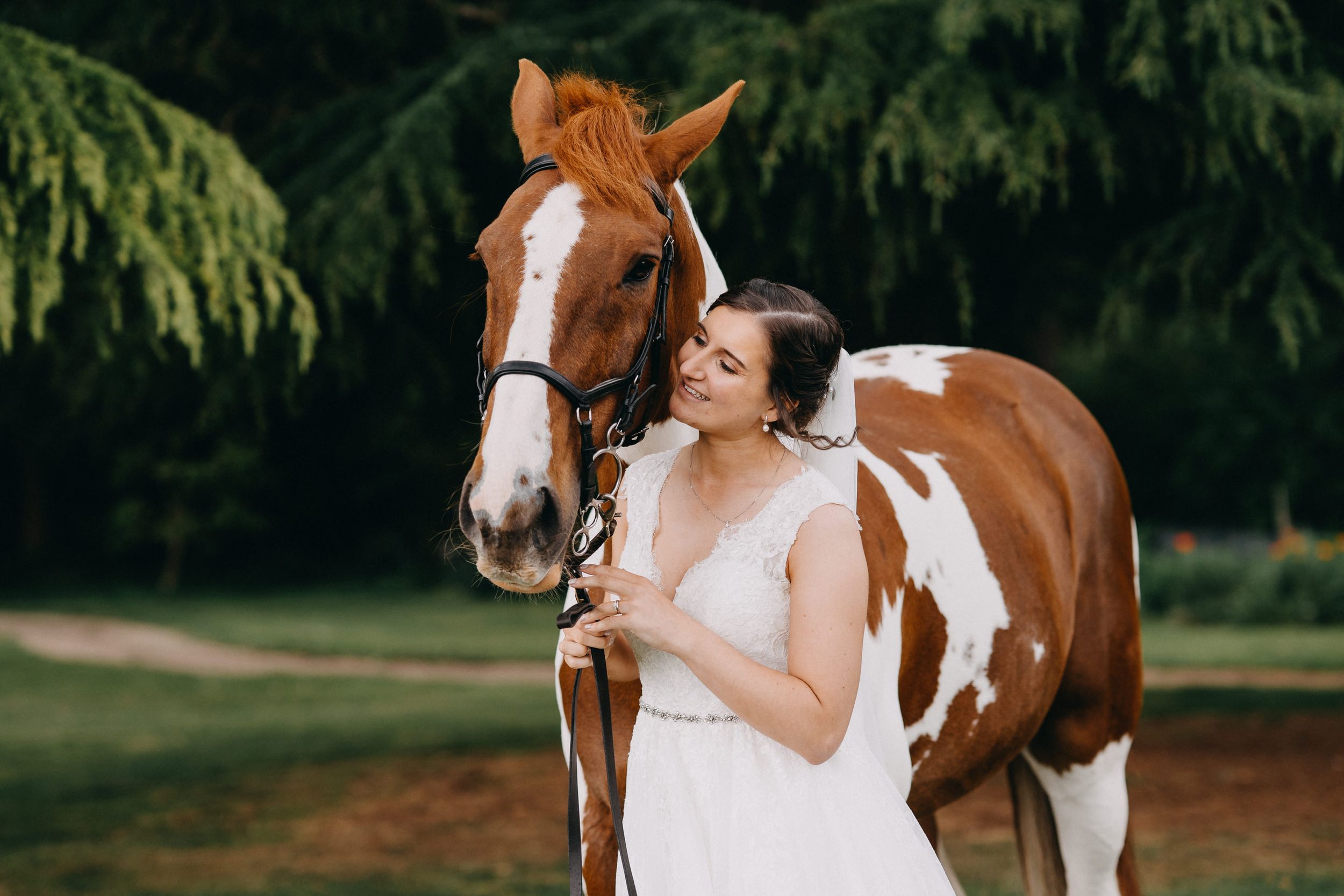 A beautiful brown and white paint horse poses with someone in a flowing white wedding dress against a natural backdrop.