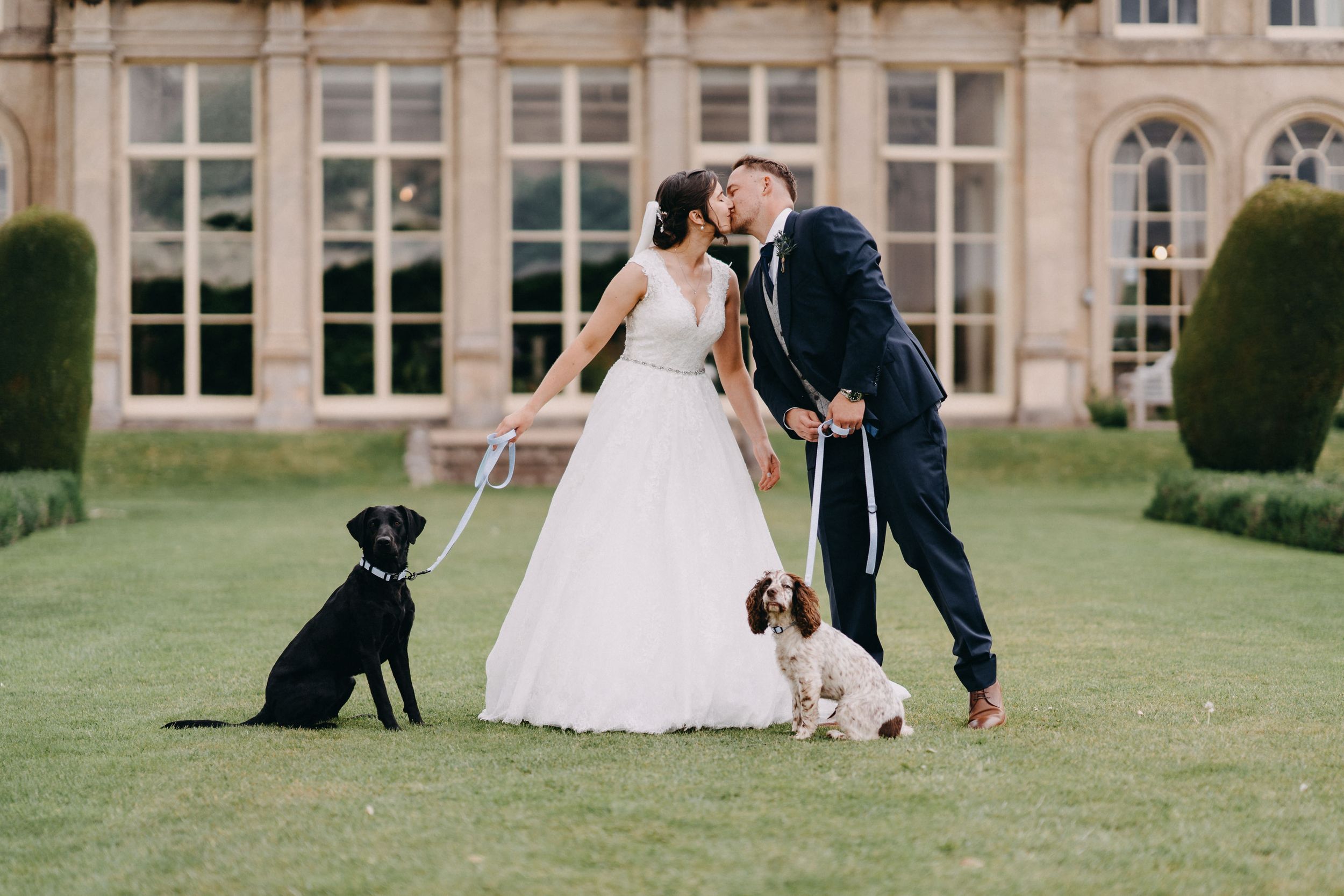 A couple shares a romantic moment on a manicured lawn with their two dogs beside them outside a grand estate building.