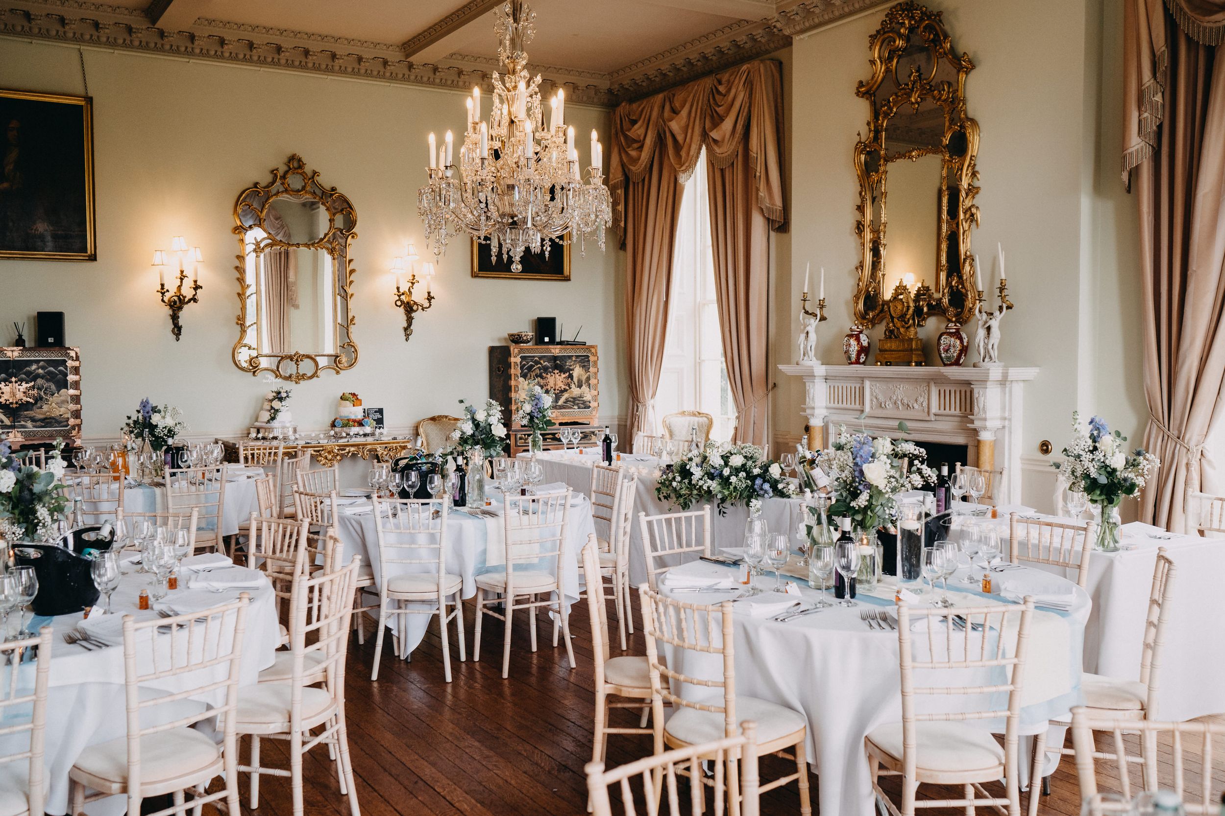 Elegant dining room at Prestwold Hall with crystal chandeliers, ornate mirrors, and white chiavari chairs set for a formal reception.