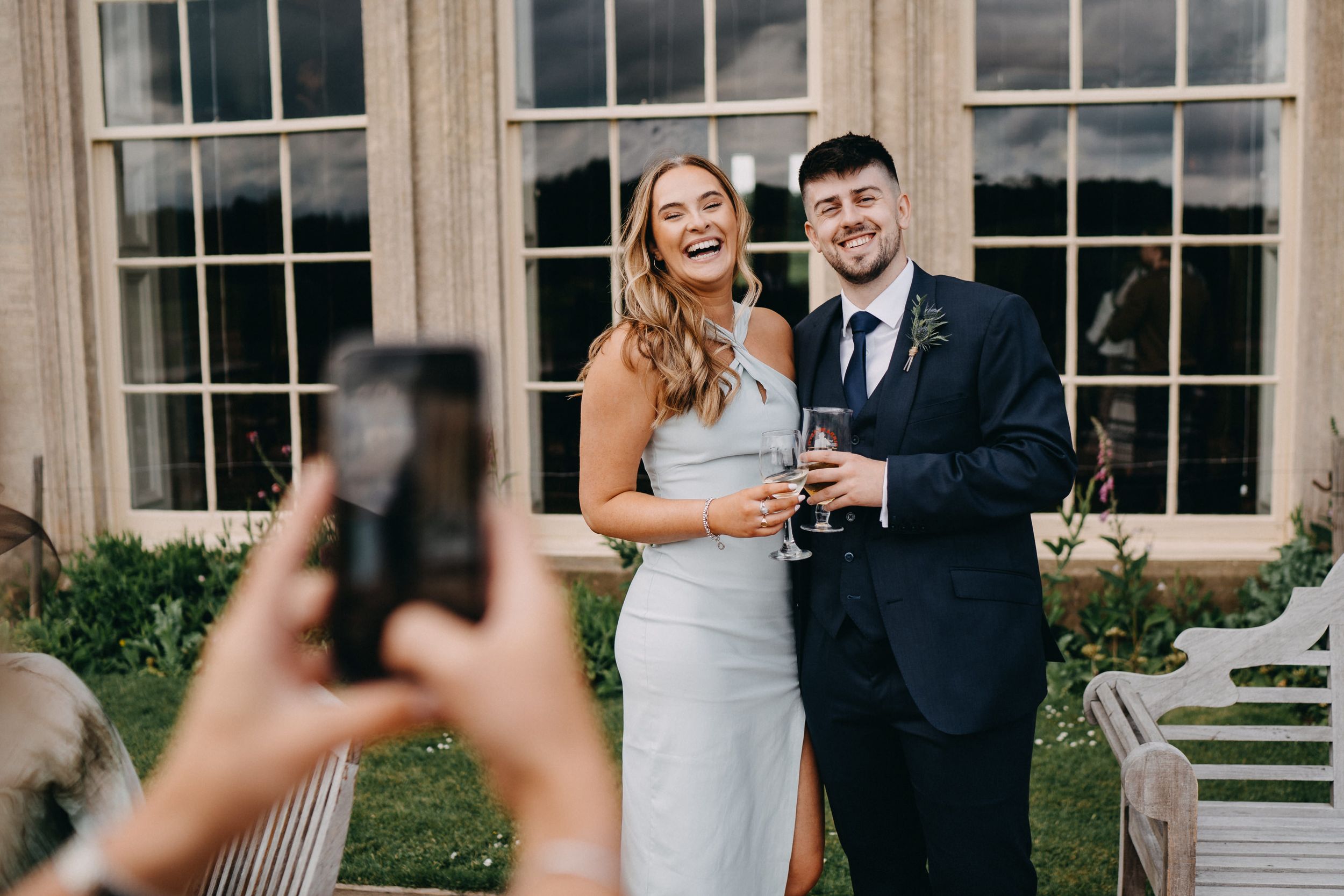 Someone takes a photo on their phone of a couple in wedding attire posing together with drinks at an elegant venue.