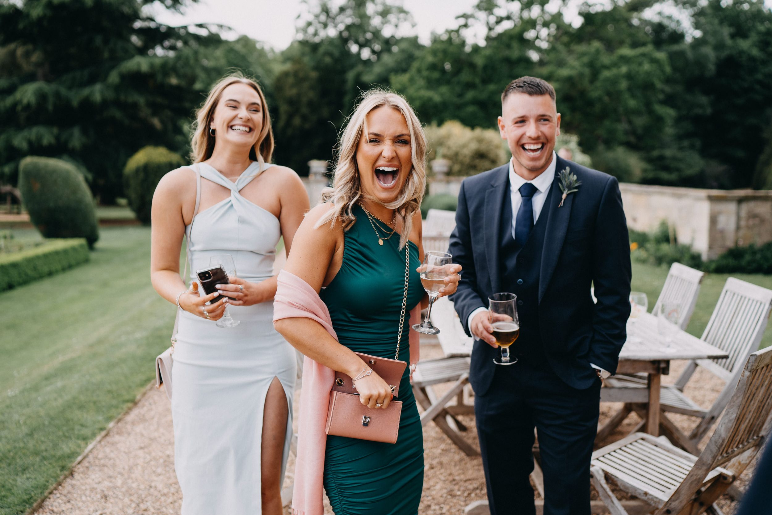 Wedding guests enjoying drinks and laughter together in a beautiful garden setting with green lawn and hedges in background.