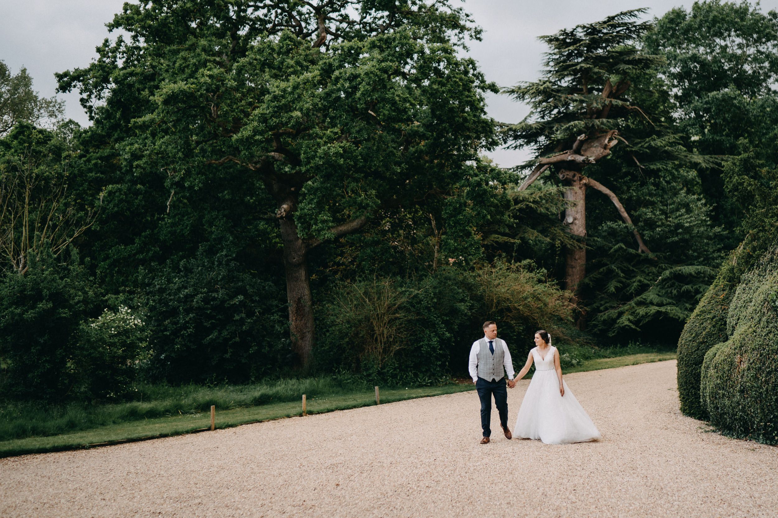 A couple strolls along a curving garden path surrounded by lush green trees and manicured hedges.