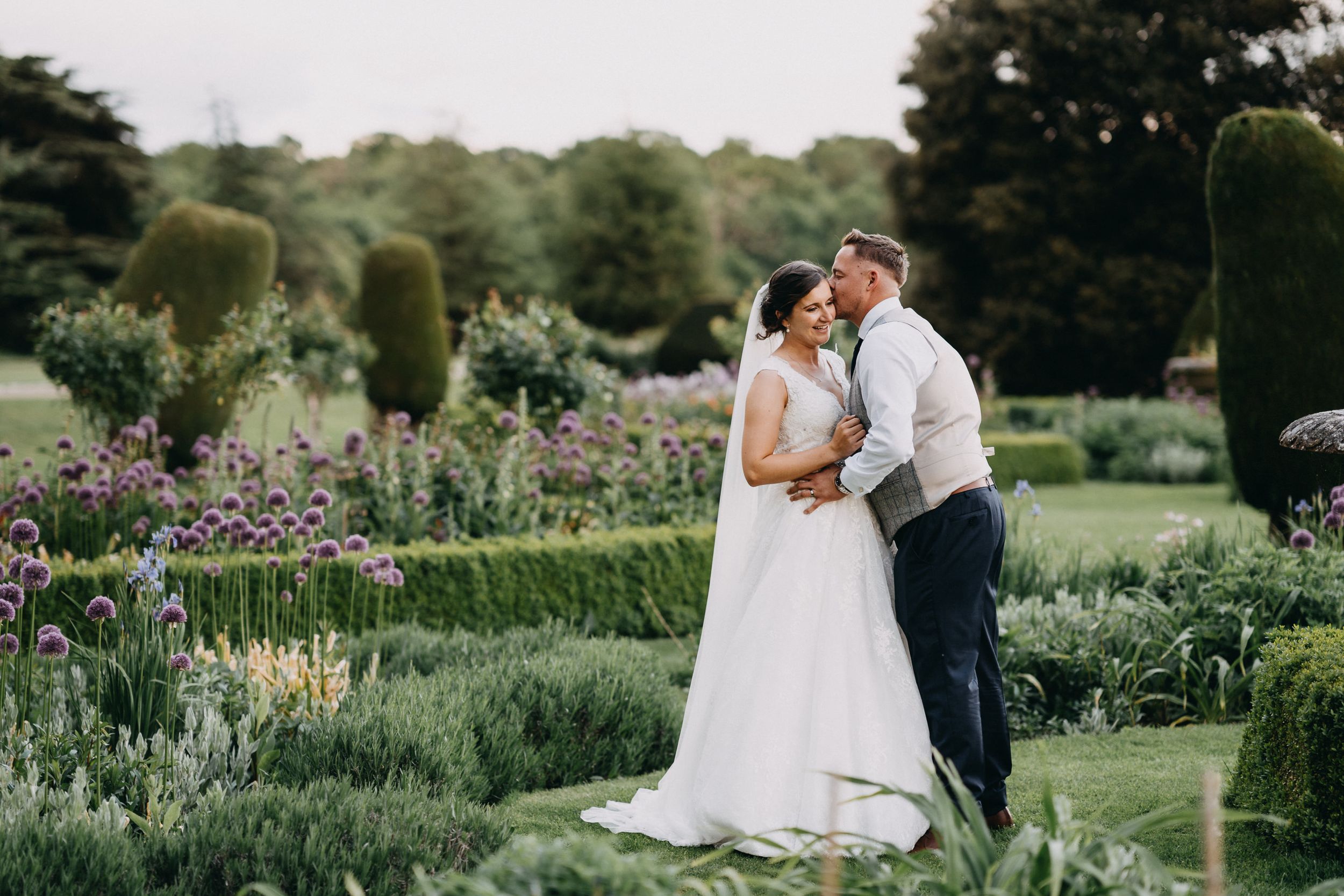 Two people share a romantic moment among blooming flowers in a formal garden with manicured hedges and pathways.