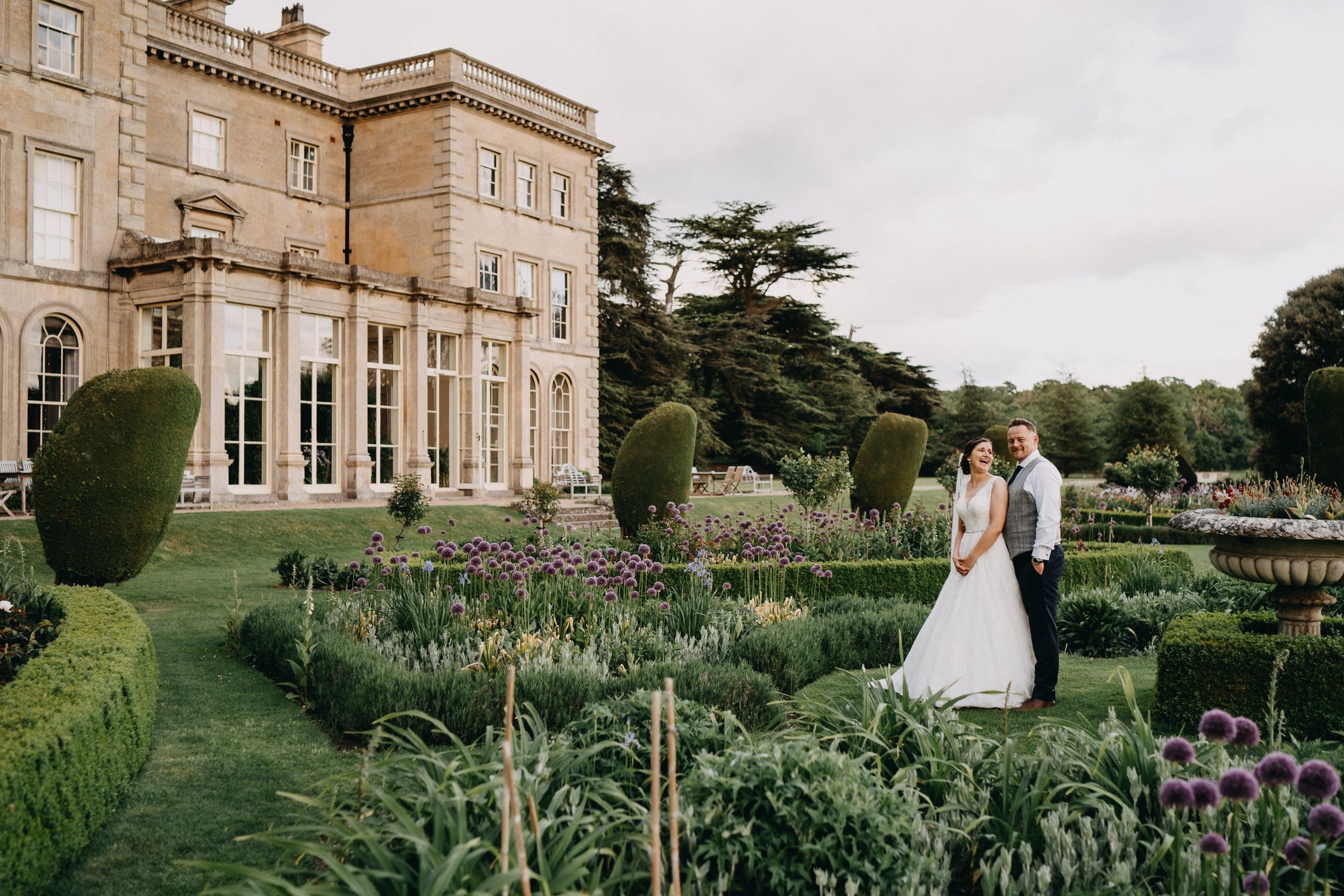 A couple stands in the manicured gardens of an elegant English manor house with stone walls and ornate hedges.