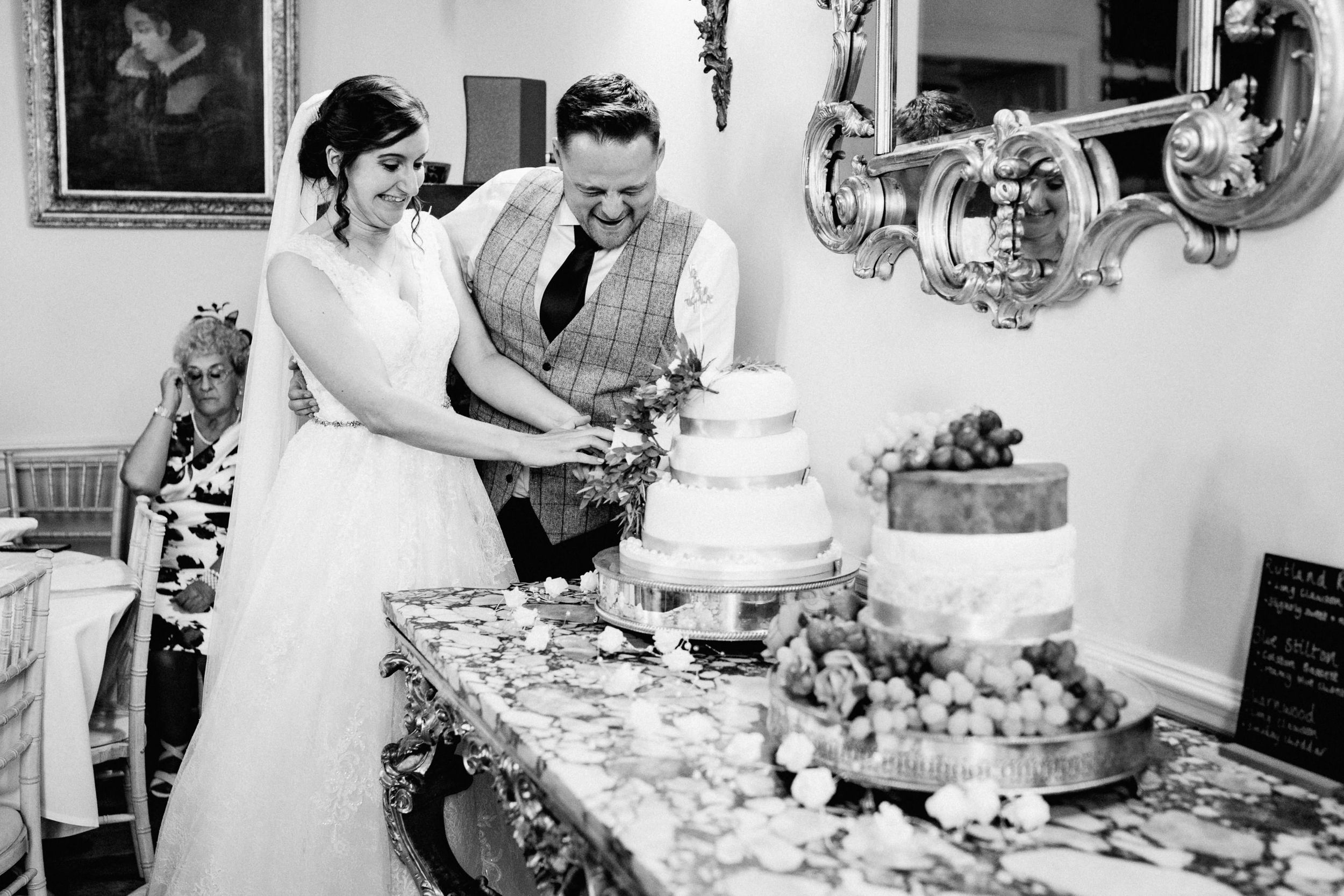 A couple cuts into their elegant multi-tiered wedding cake at an indoor reception decorated with ornate mirrors.
