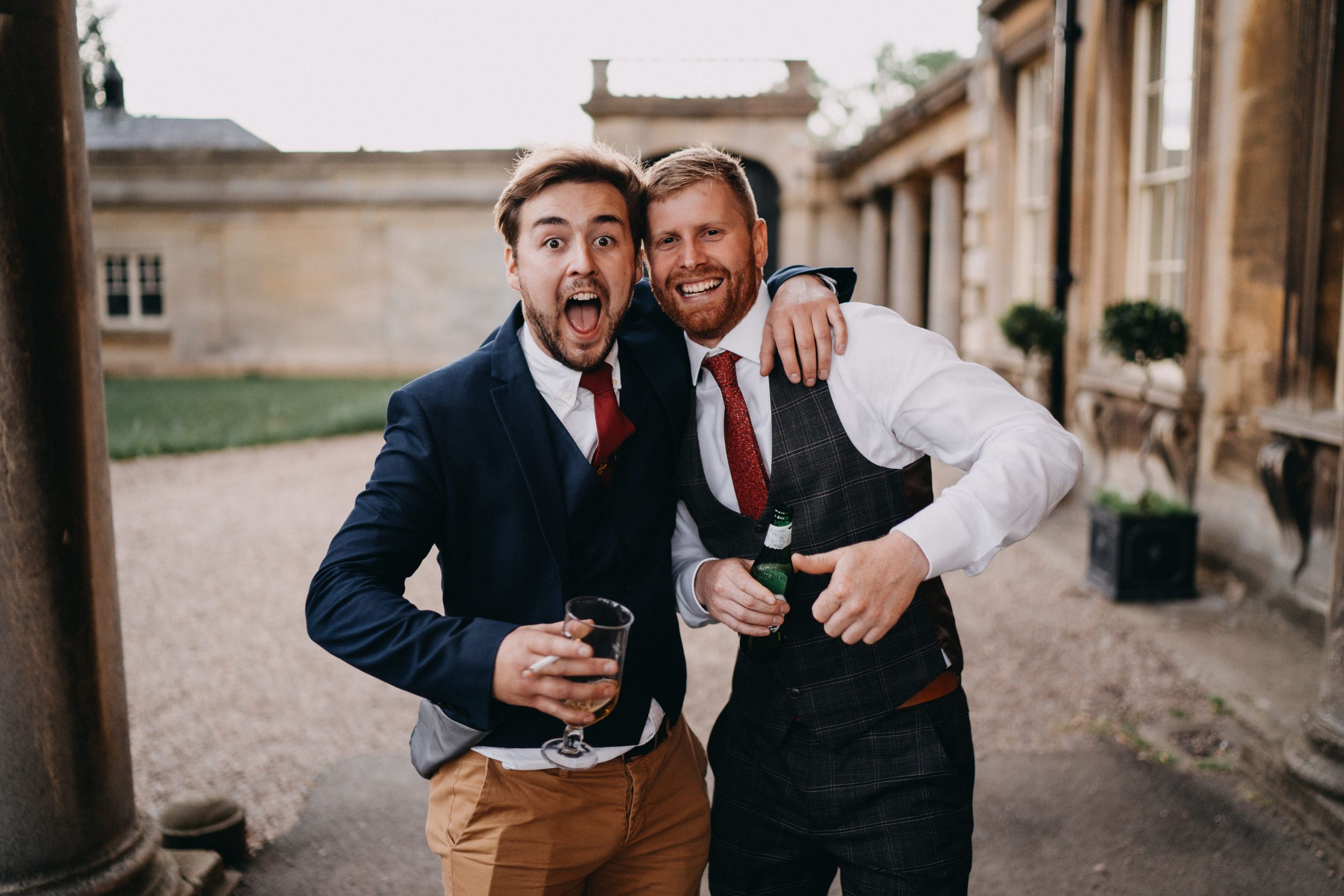 Two well-dressed friends in suits share drinks and smiles during an outdoor celebration at a stone building.