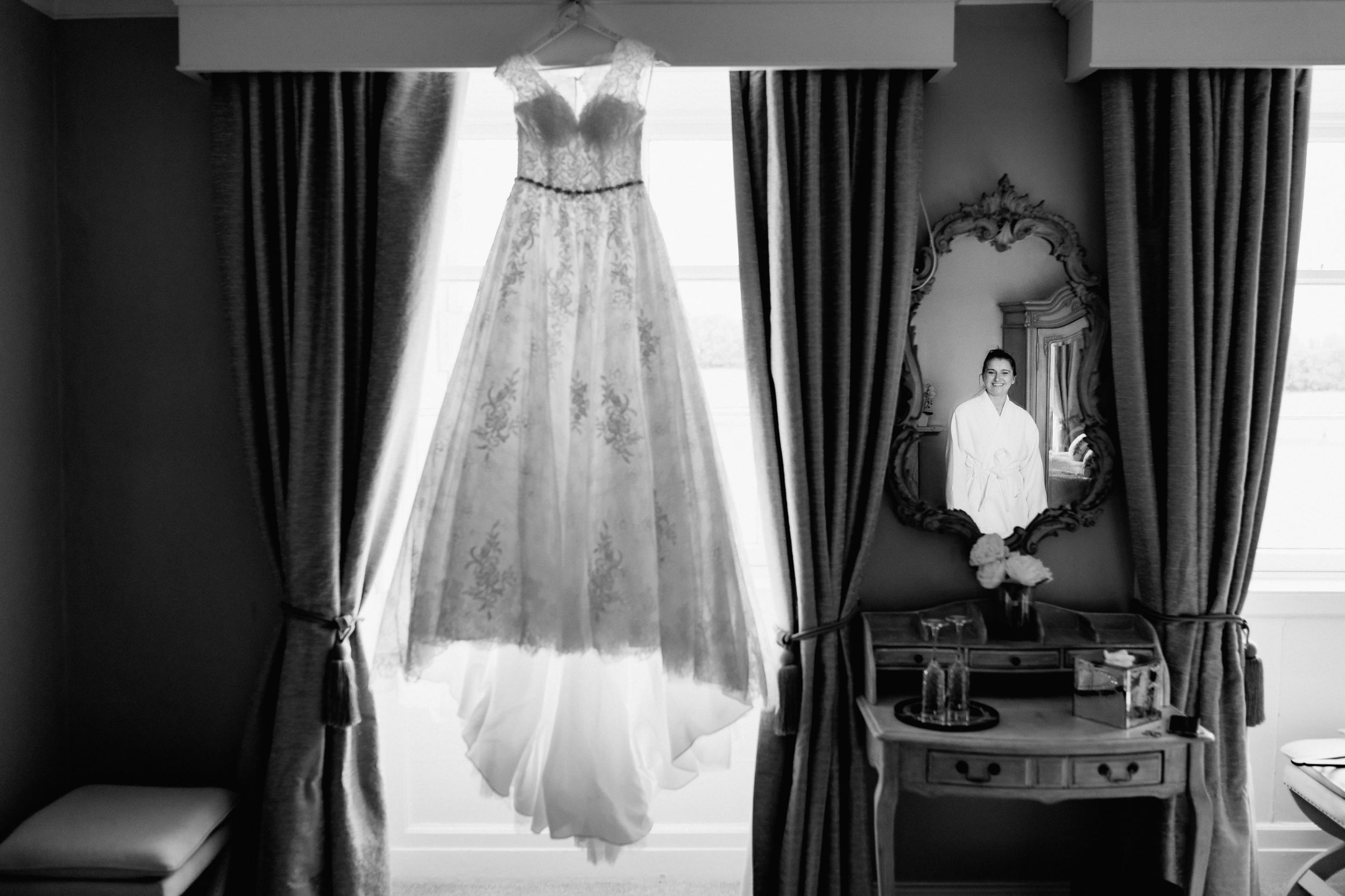 Wedding dress hanging in window light between dark curtains with antique mirror and vanity visible.