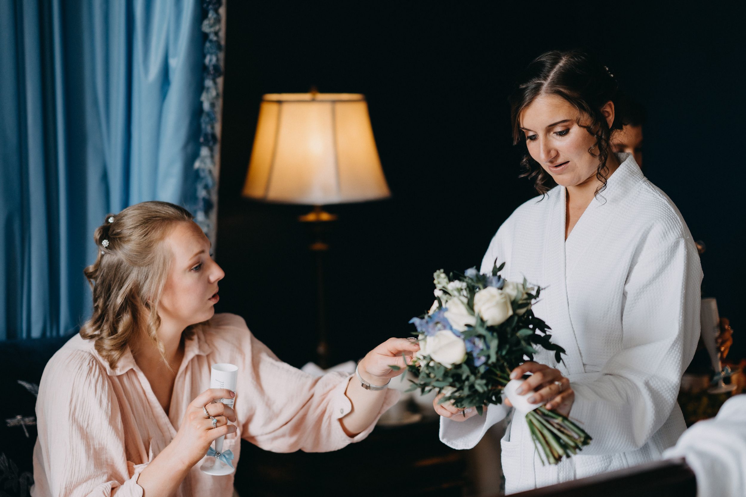 Two people in robes share a tender moment while looking at a white floral bouquet in a dimly lit room.