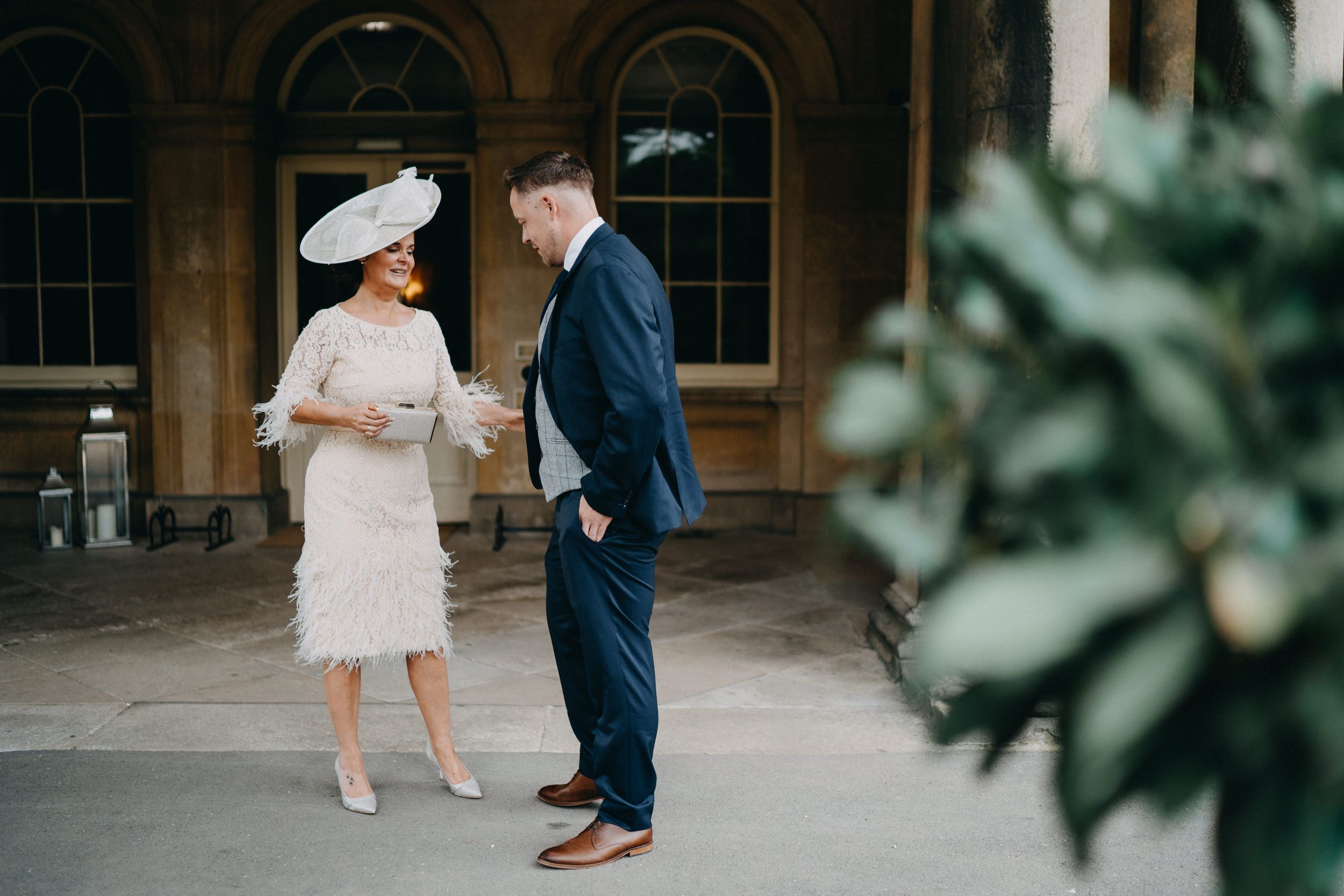 Two people in formal attire share a moment together at an elegant venue with arched windows and greenery.