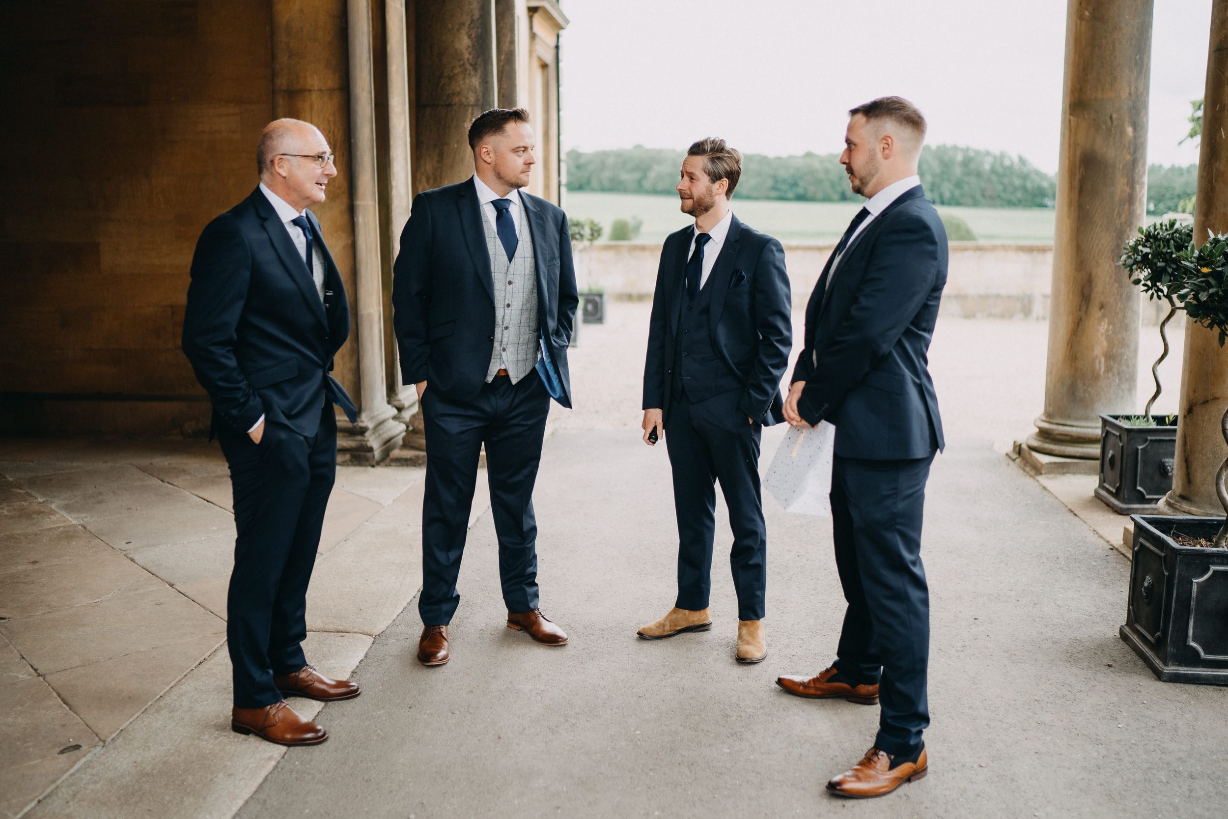 Four men in dark suits and dress shoes chat casually under stone archway columns.